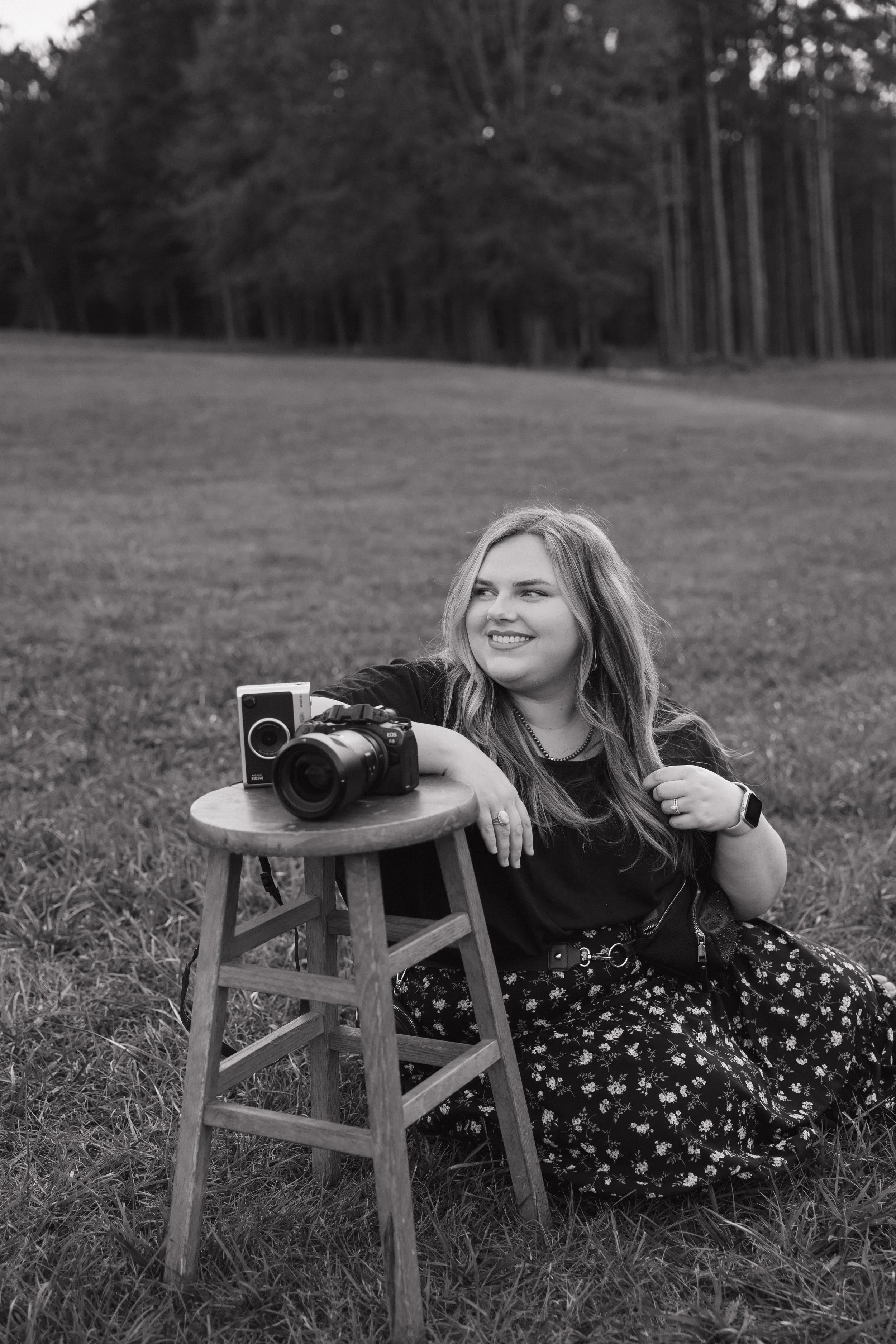 A woman sitting on grass outdoors with a camera on a small stool, smiling and looking to the side, in black and white.