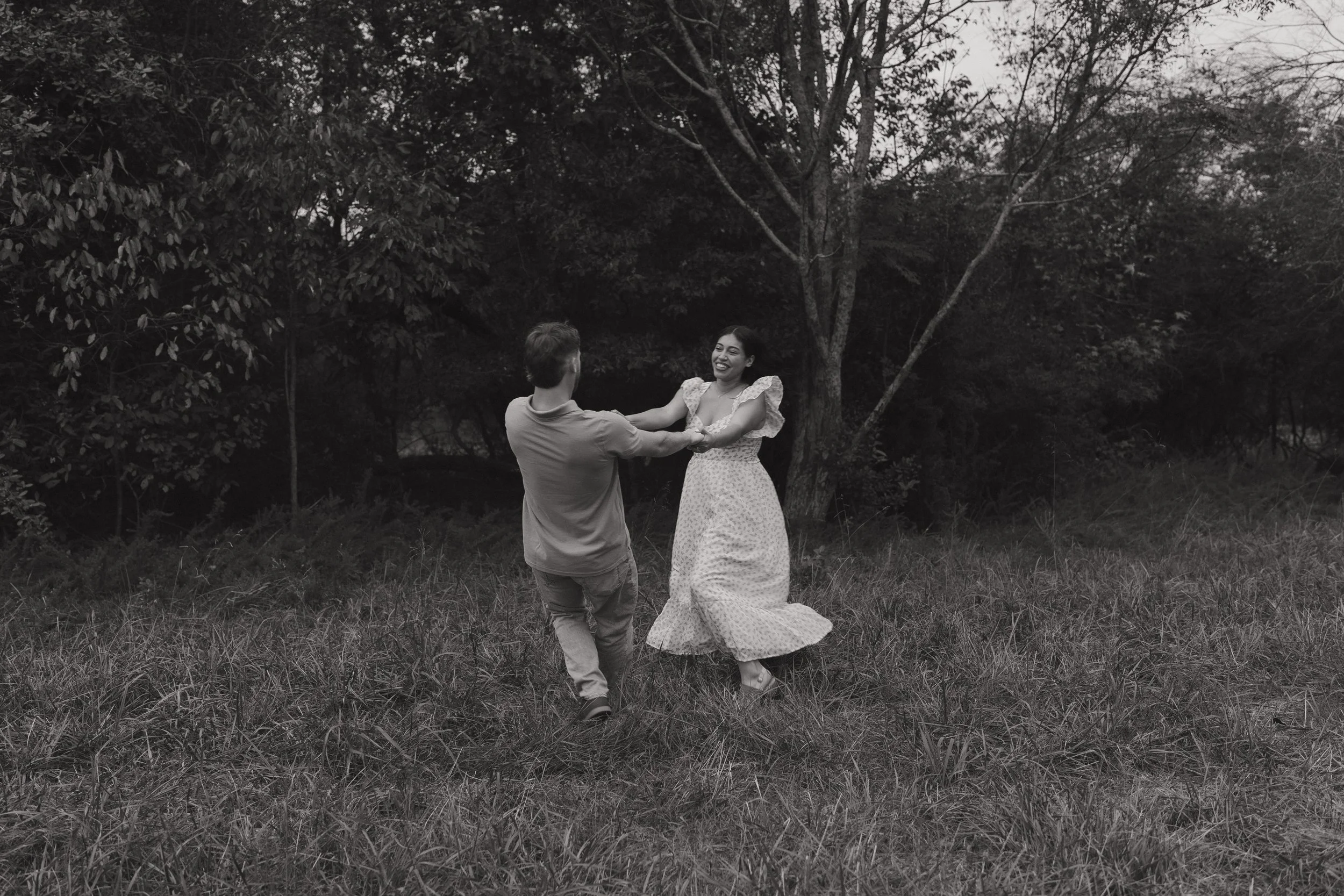 A black and white photo of a couple dancing outdoors in a grassy area with trees in the background, smiling and holding hands.