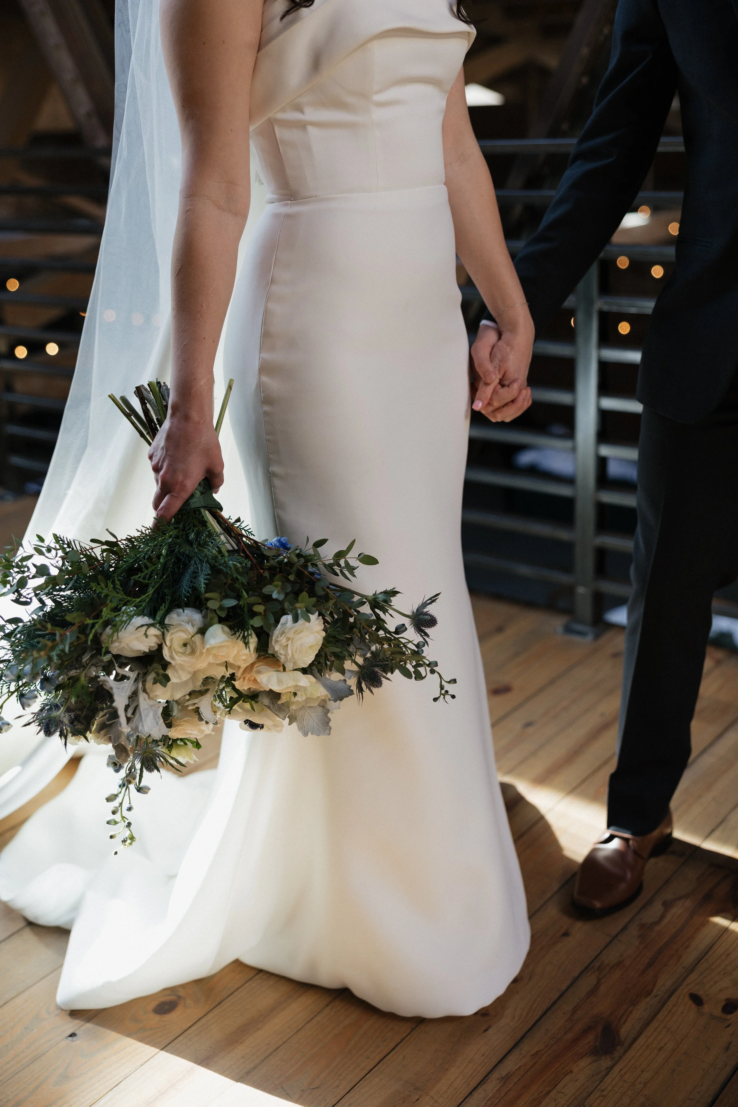 Close-up of a bride in a white wedding dress holding a bouquet of white and green flowers, standing next to a groom in a black suit, at a wedding ceremony.