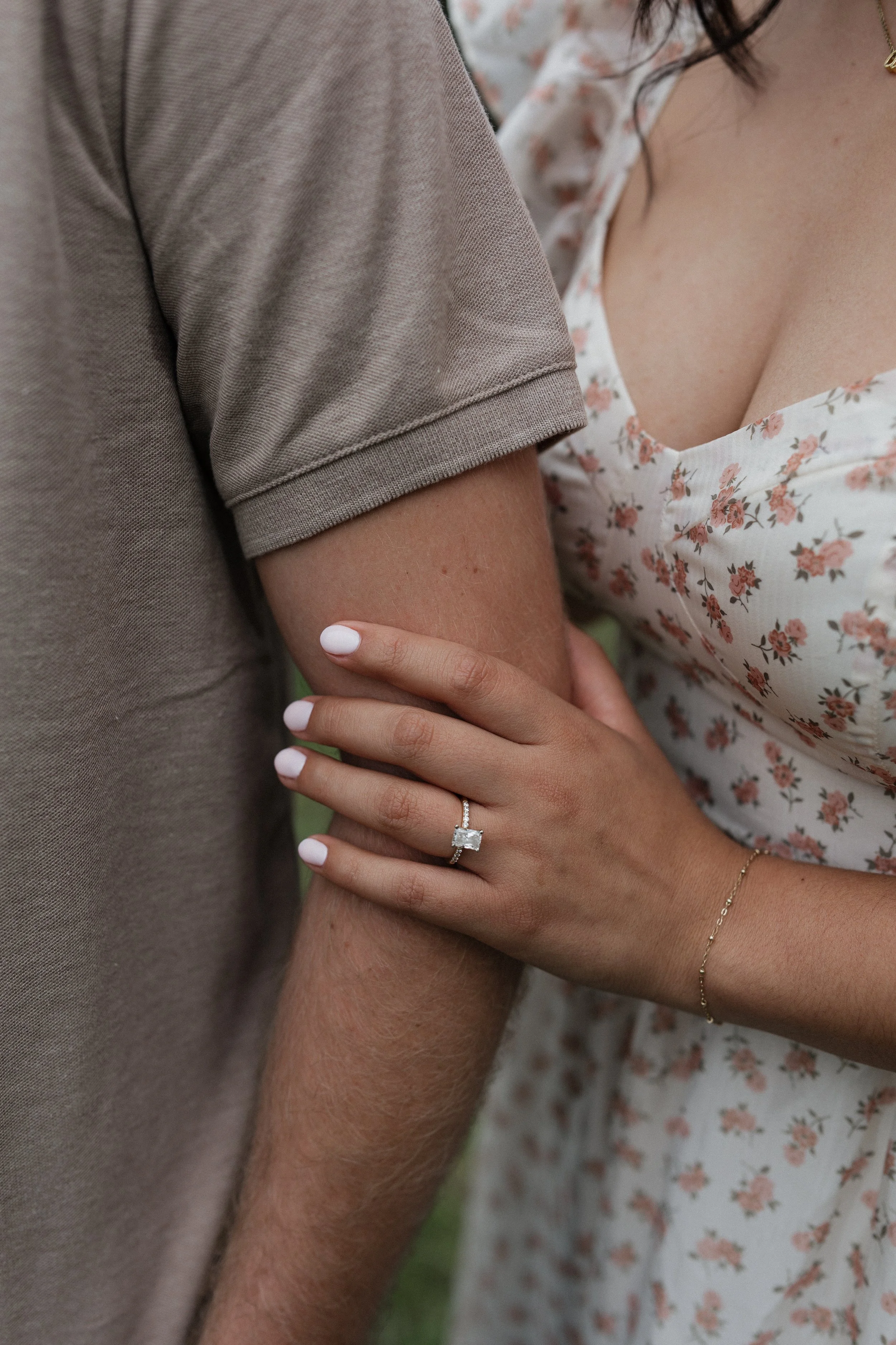 A woman wearing a floral dress holding a man's arm, displaying an engagement ring with a large rectangular diamond on her finger.