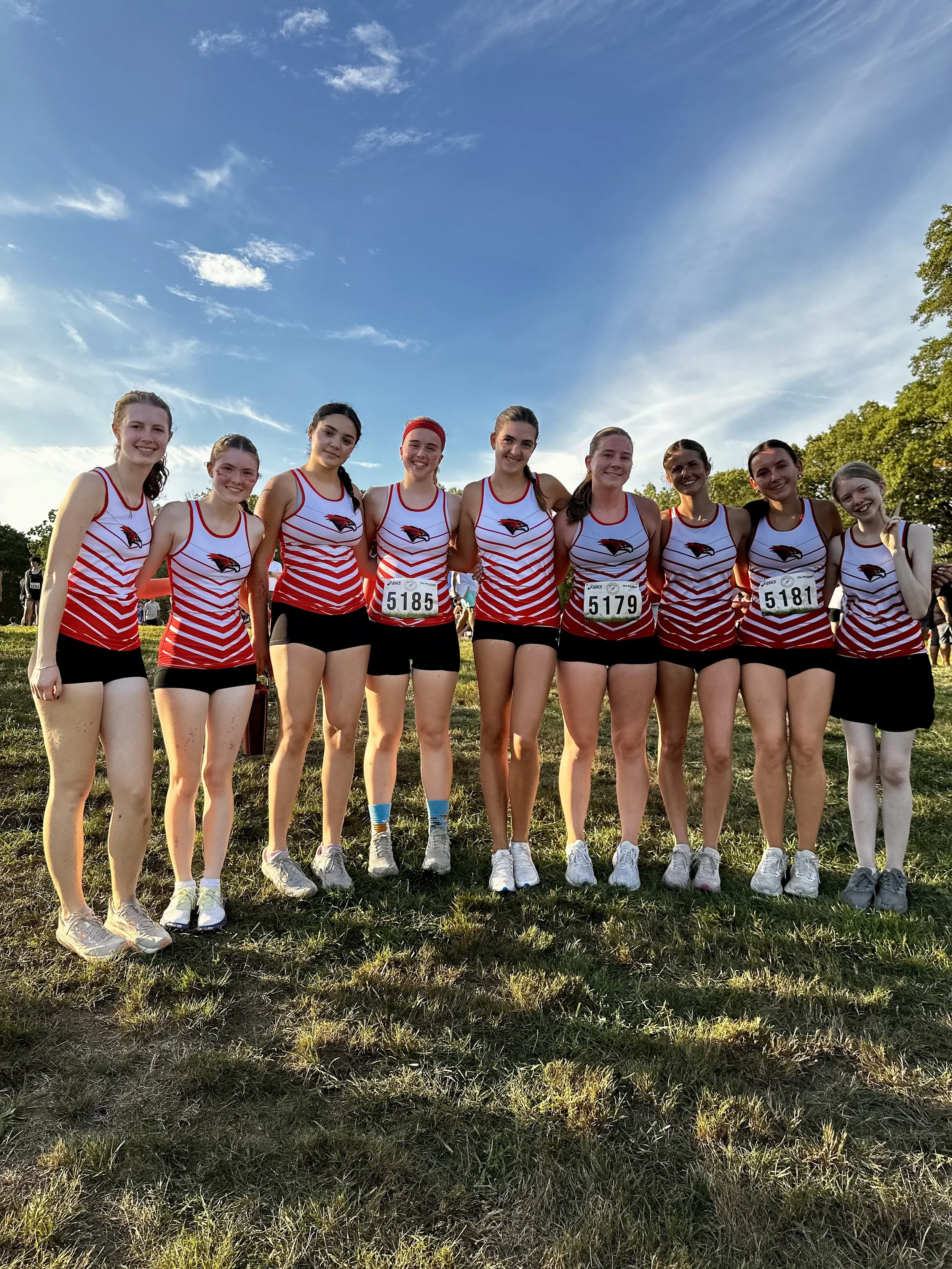 Group of high school cross country runners starting a race on a grassy field with a playground in the background, surrounded by trees with autumn leaves.