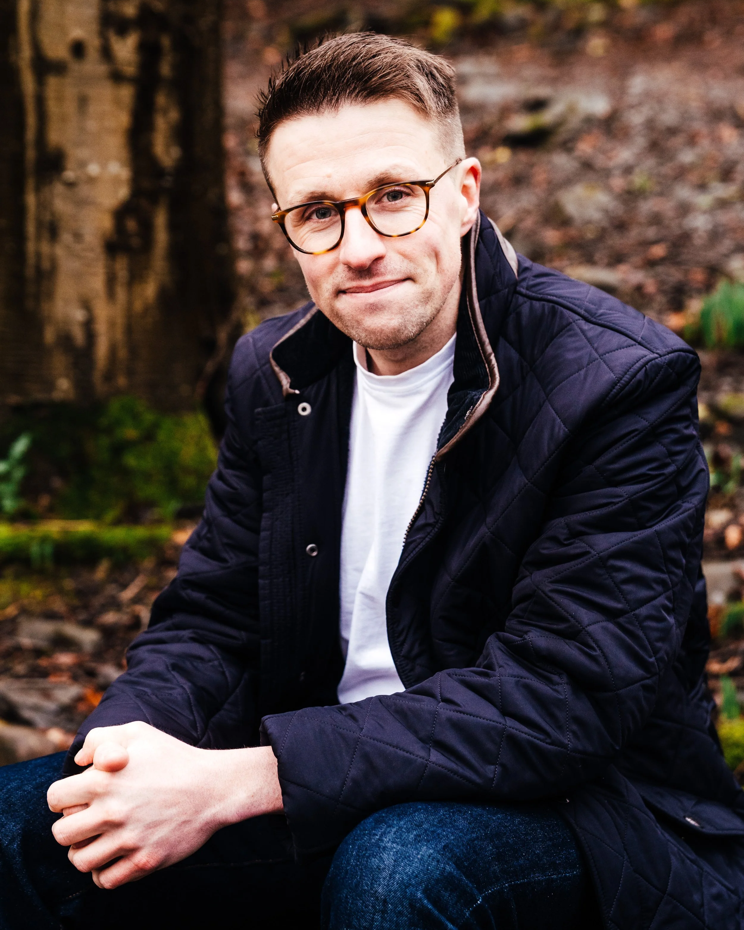 Andy is wearing glasses and a navy quilted jacket sitting outdoors on a forest floor.