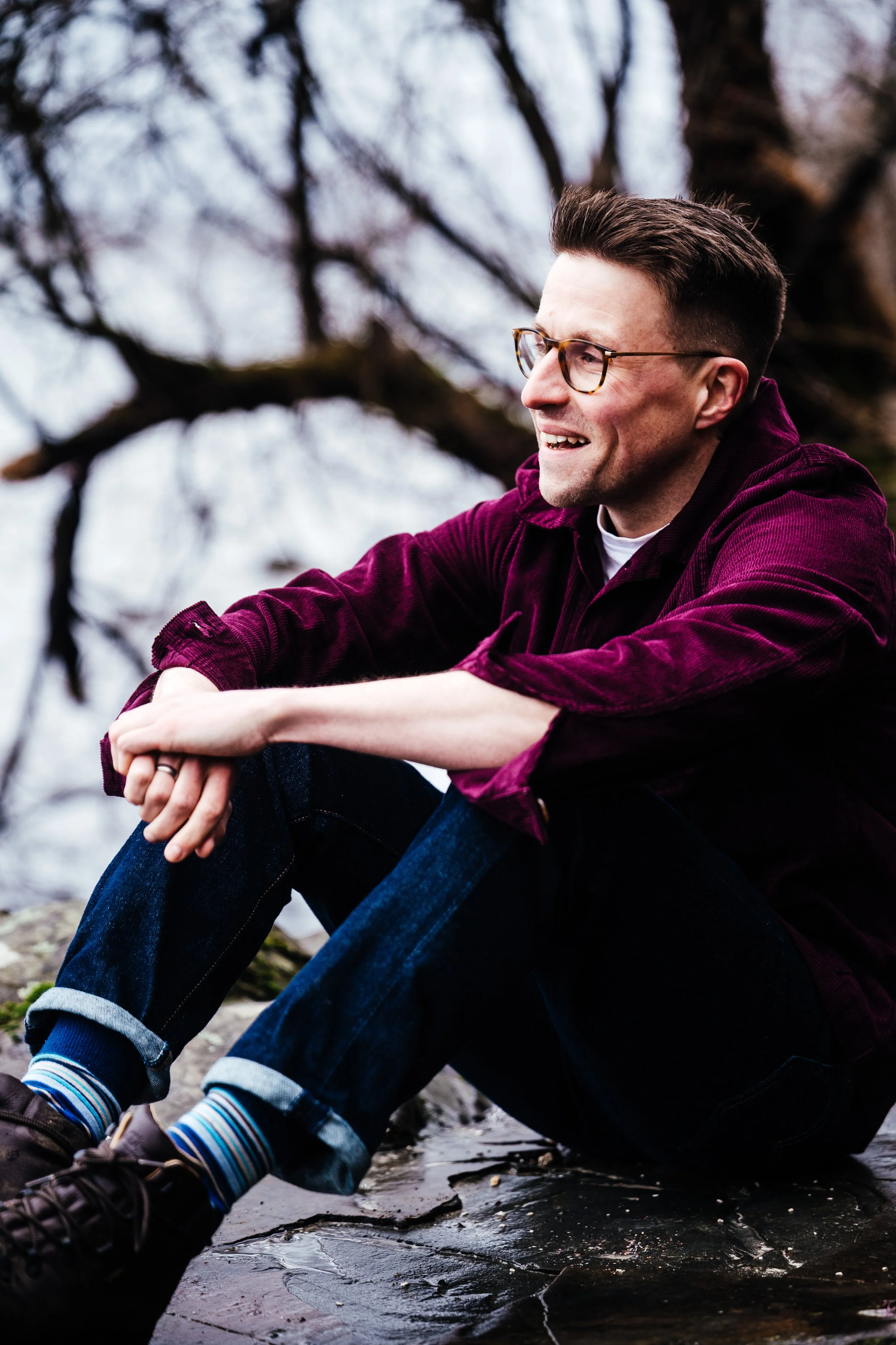 A picture of Andy, sat on a fallen tree in a forest, looking into the distance and smiling.