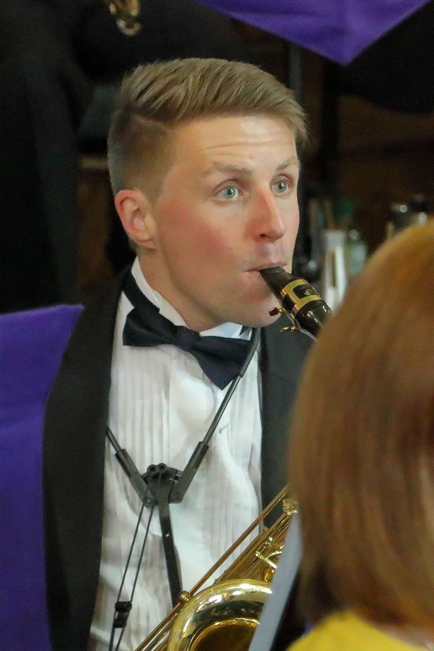 Andy hunt is wearing a white shirt and a black bow tie, sitting down in a band playing baritone saxophone.