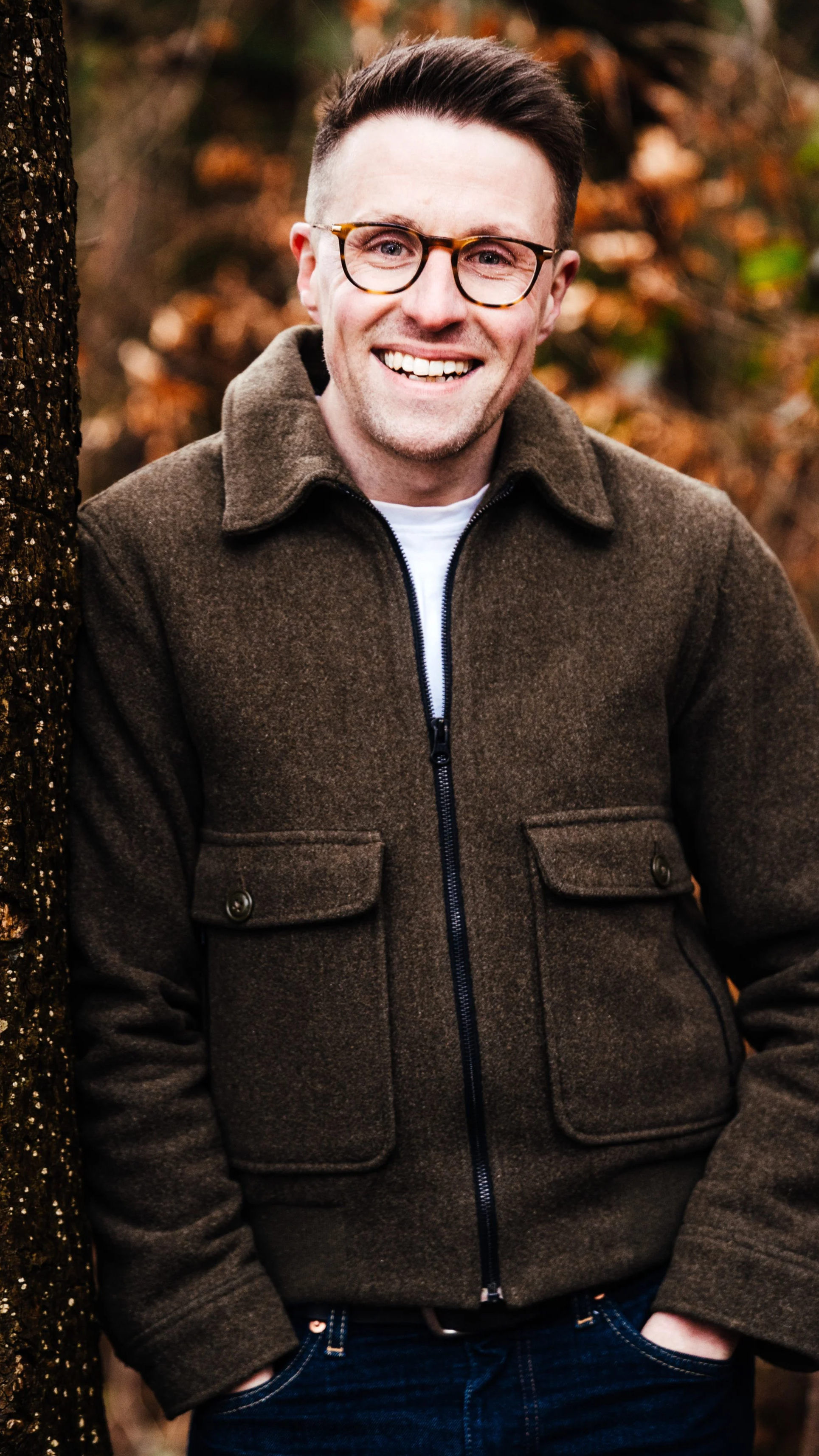 Andy has short dark hair and glasses leaning against a tree, smiling in an autumn forest, wearing a brown jacket and blue jeans.