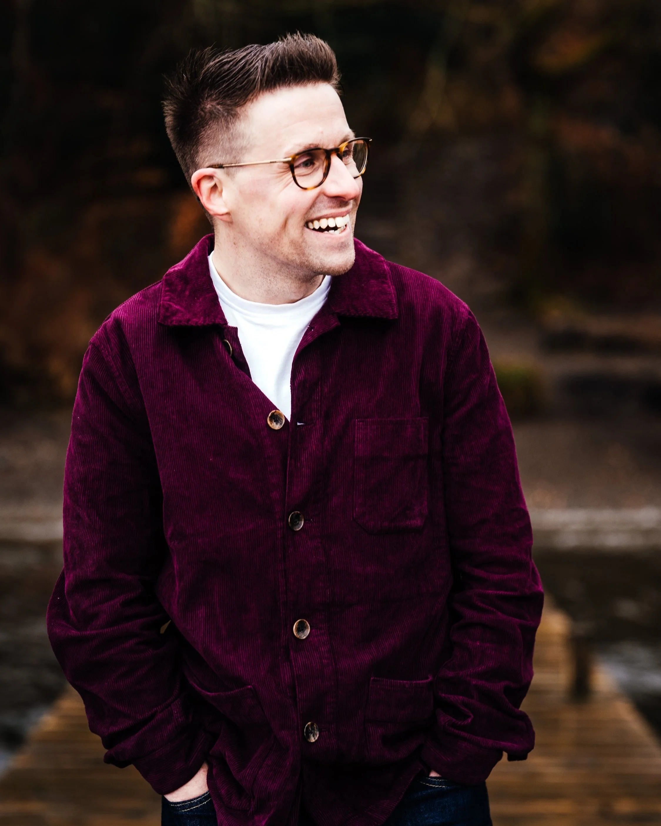Andy Hunt is wearing glasses, a purple jacket, and a white shirt is smiling and looking to the side, standing in an outdoor setting.