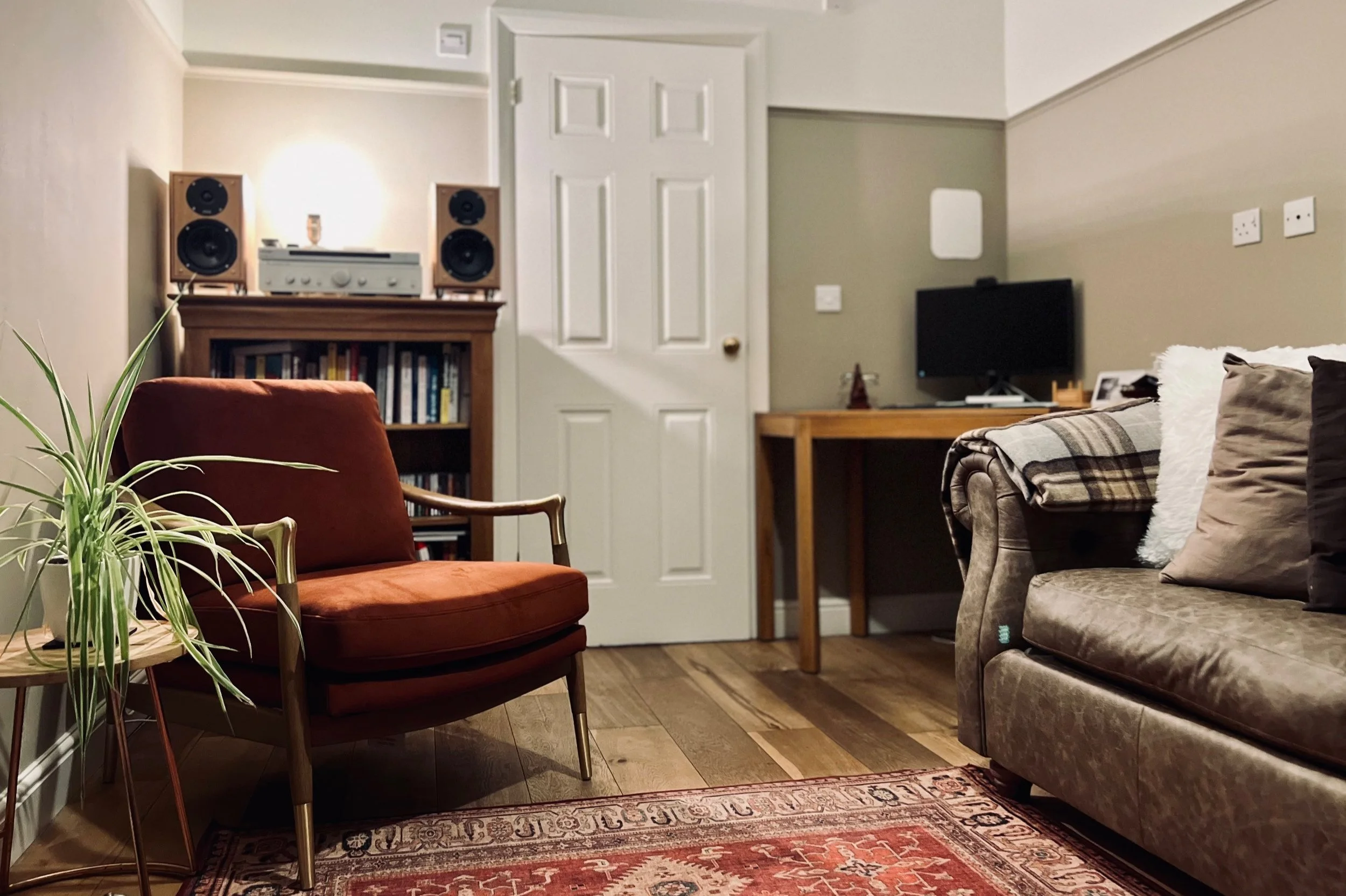 Cozy therapy room with a brown leather sofa, orange armchair, indoor plant, wooden bookshelf, stereo system, and computer desk with monitor.
