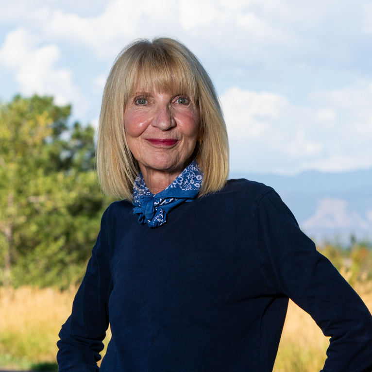 Older woman with blonde hair wearing a navy blue top and a blue bandana, standing outdoors with trees and mountains in the background.