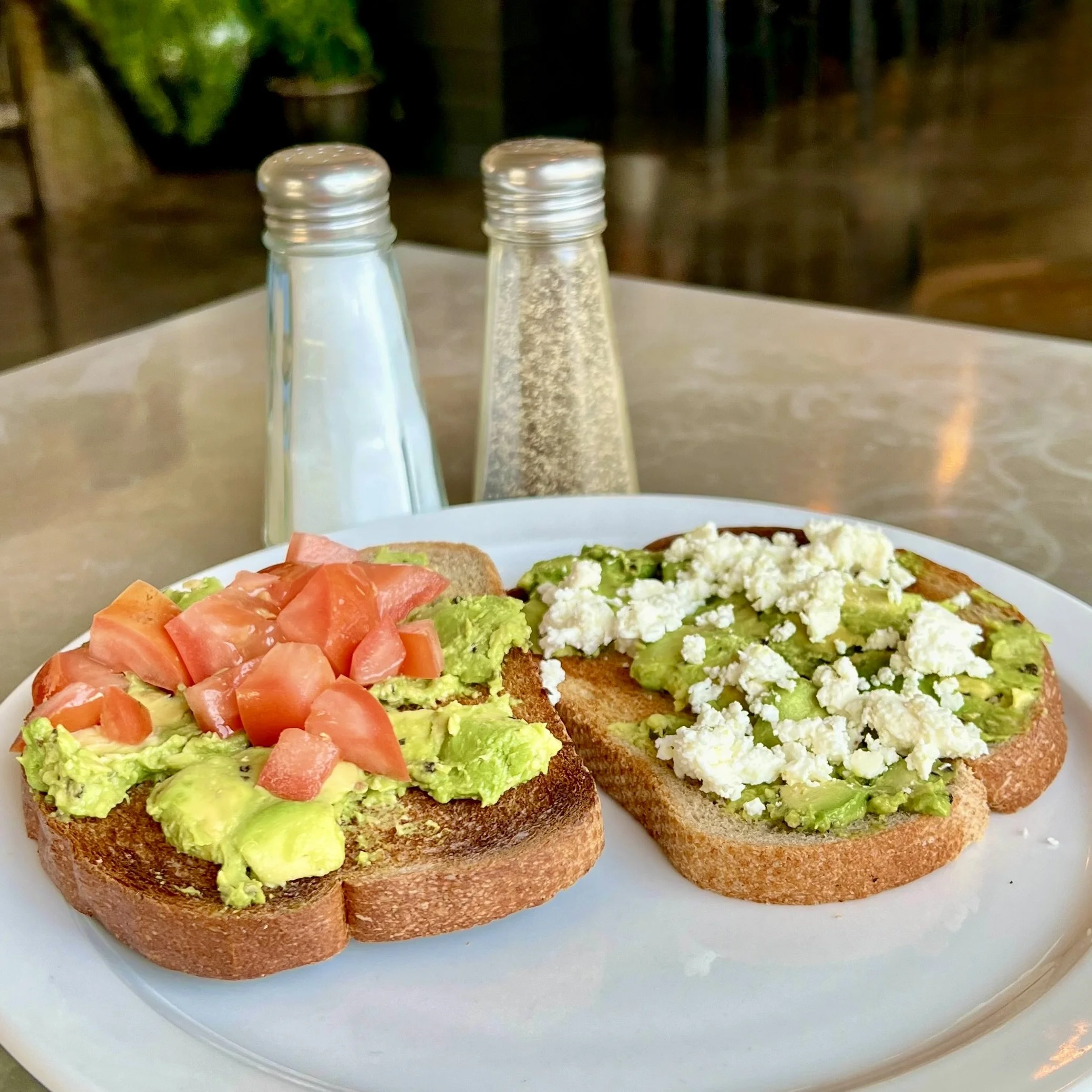 Two slices of toasted bread topped with guacamole, tomatoes, and crumbled cheese on a white plate, with salt and pepper shakers in the background.