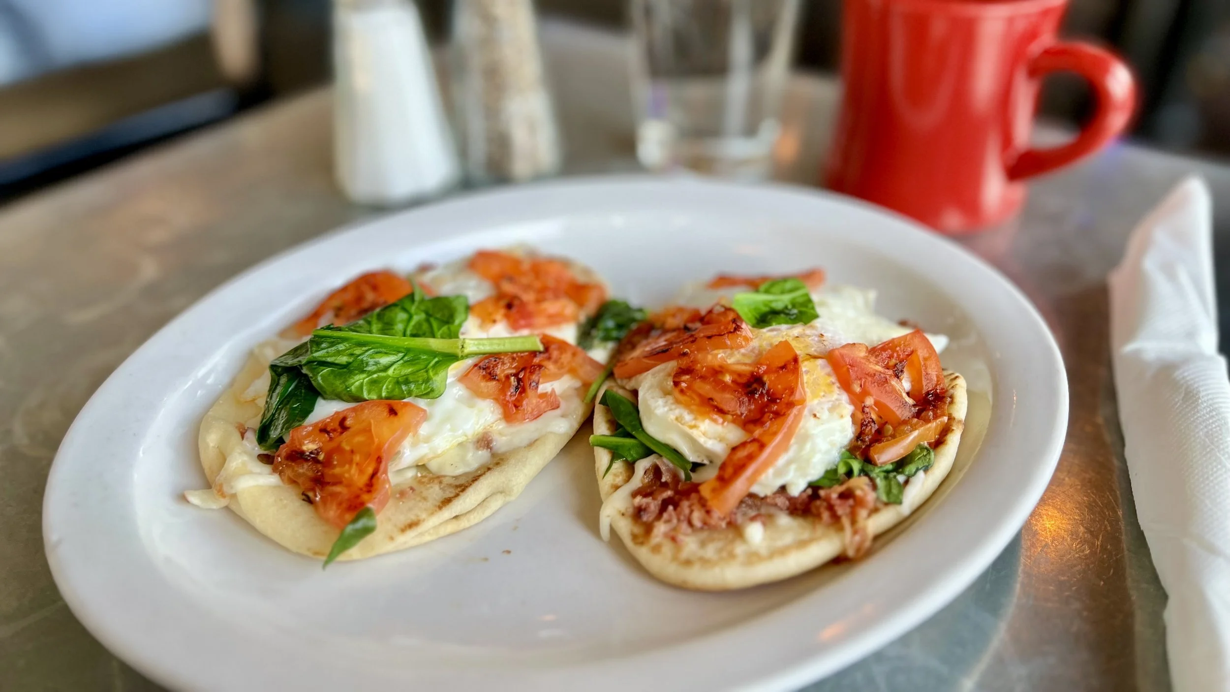 Two breakfast pizzas topped with eggs, tomatoes, green leafy vegetables, and cheese on a white plate