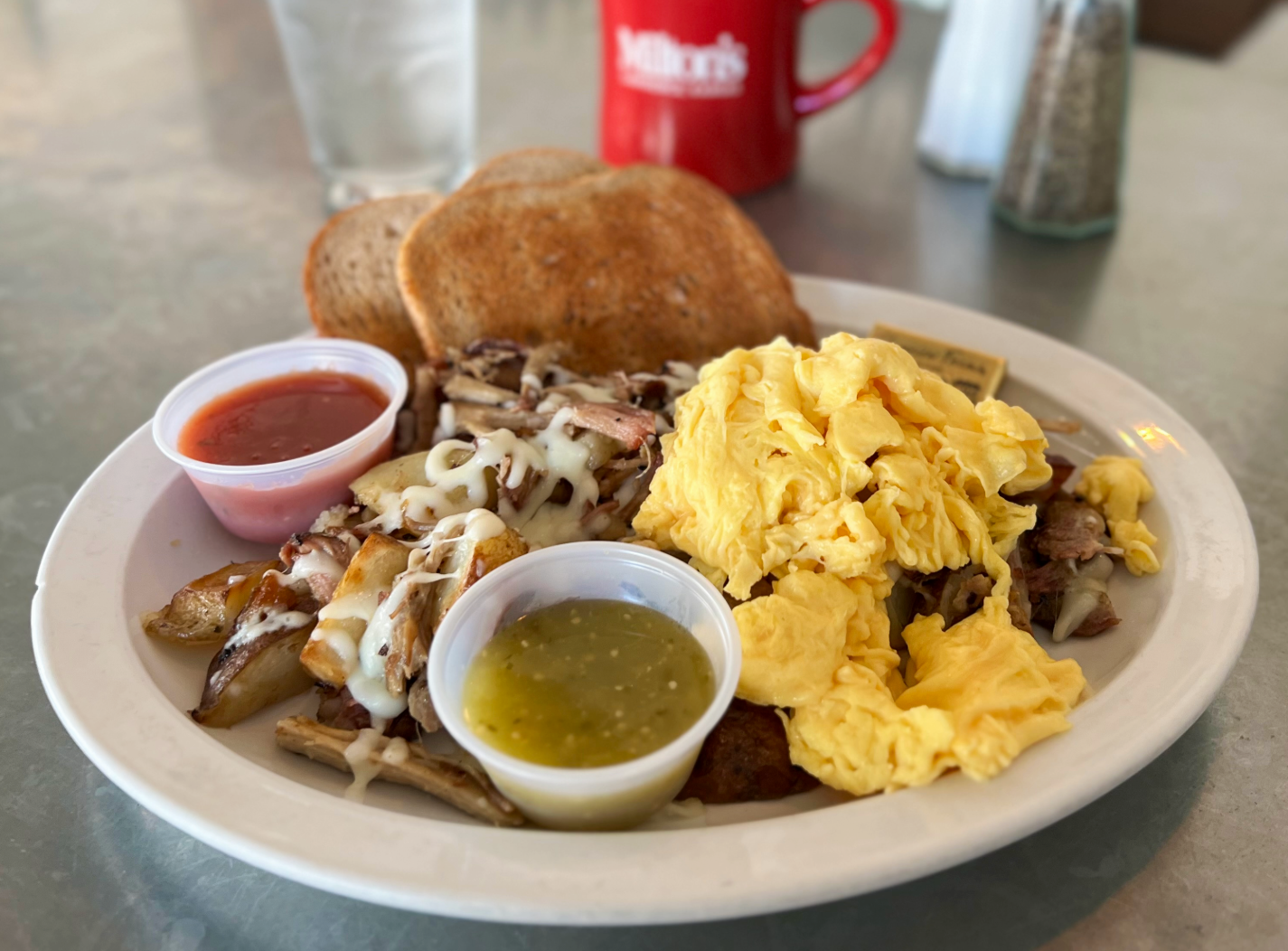 A breakfast plate with scrambled eggs, toast, shredded meat with cheese, roasted potatoes with cheese, and two small cups of salsa and green sauce.