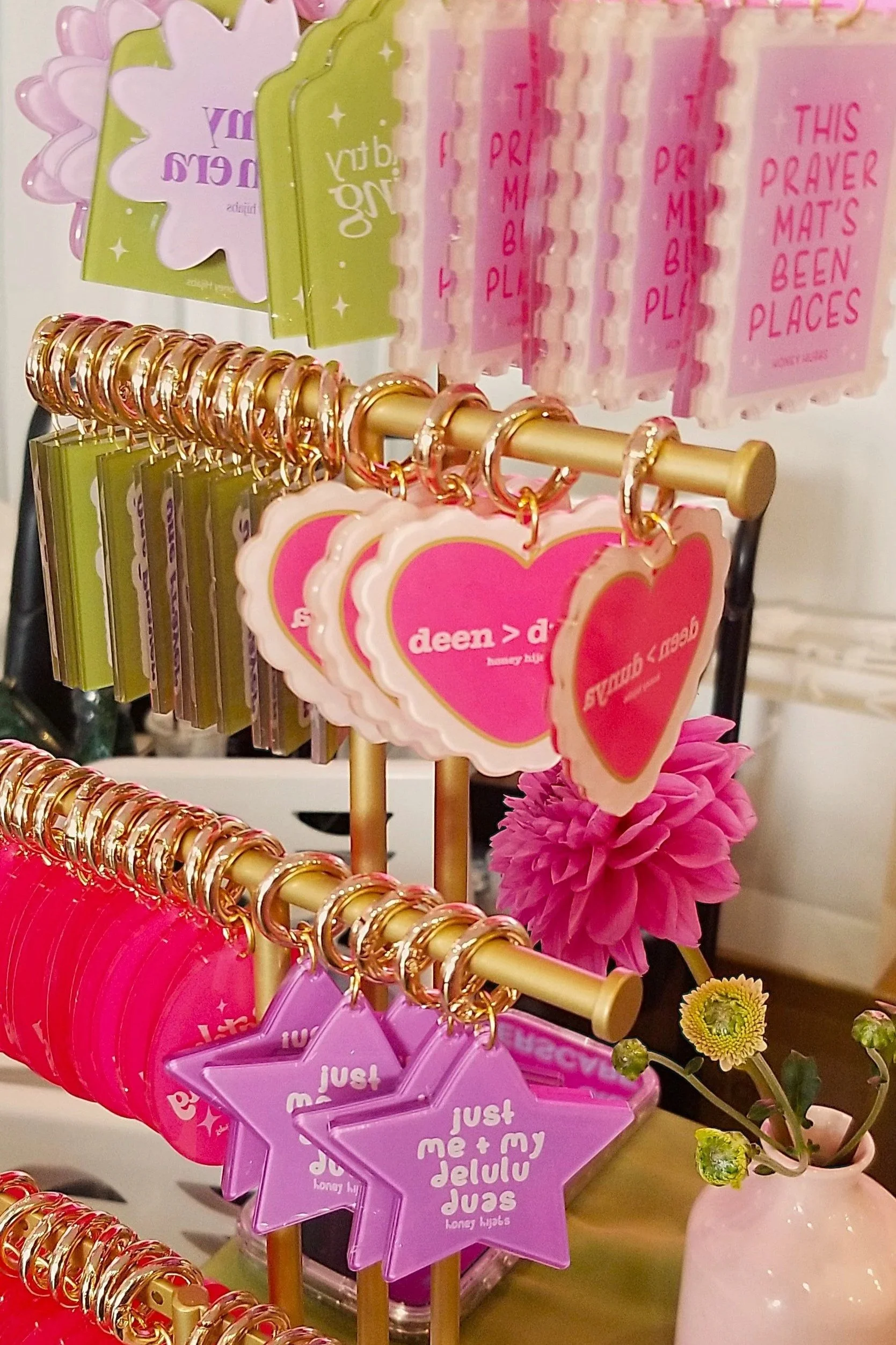 Display of pink, green, purple, and white keychains with various shapes and messages, including hearts, stars, and phrases like 'just me + my delulu duas' and 'this prayer mat's been places,' on a green table.