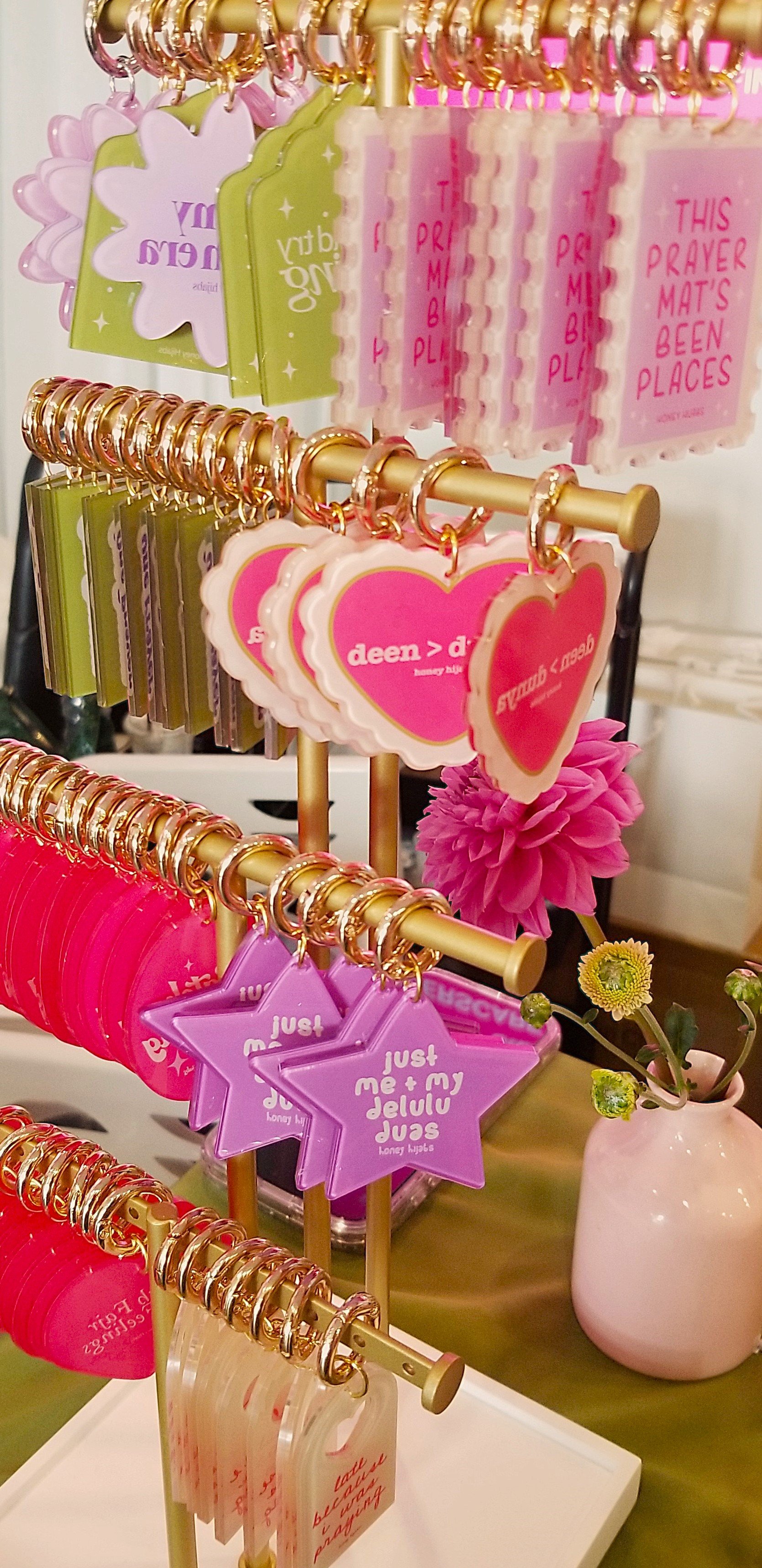 Display of pink, green, purple, and white keychains with various shapes and messages, including hearts, stars, and phrases like 'just me + my delulu duas' and 'this prayer mat's been places,' on a green table.