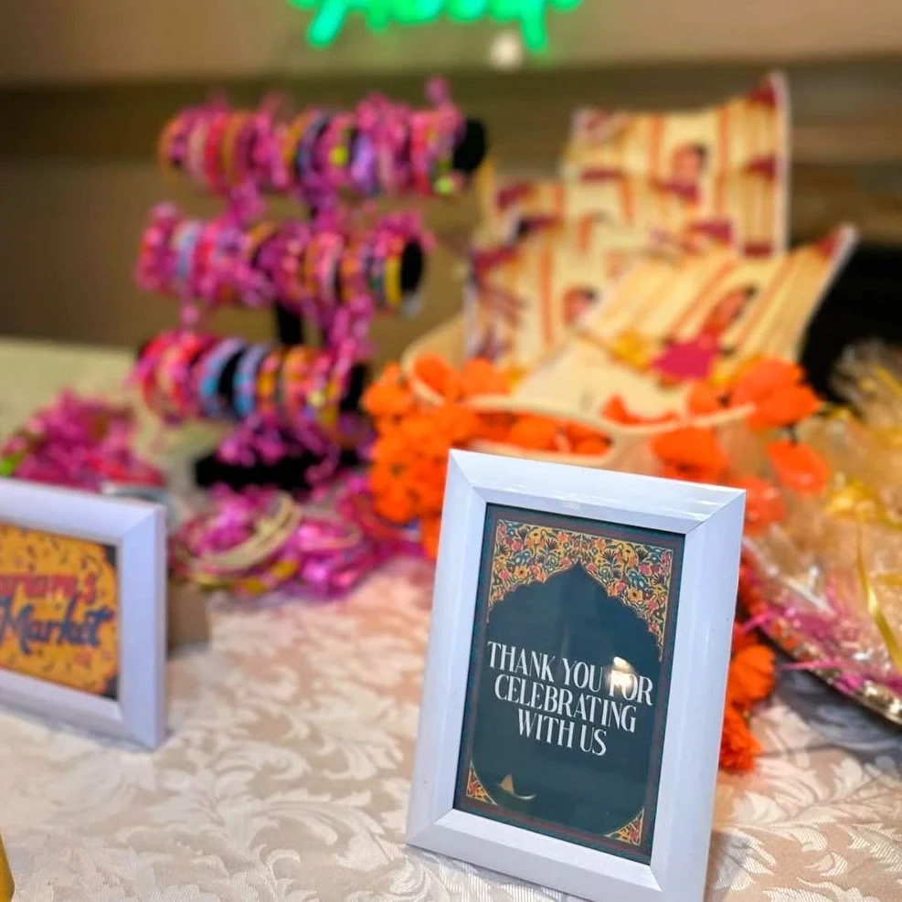 Decorative table setup with a white-framed thank you sign, colourful wrapped candies on a candy tower, flowers, and other treats, at a mehndi wedding event.