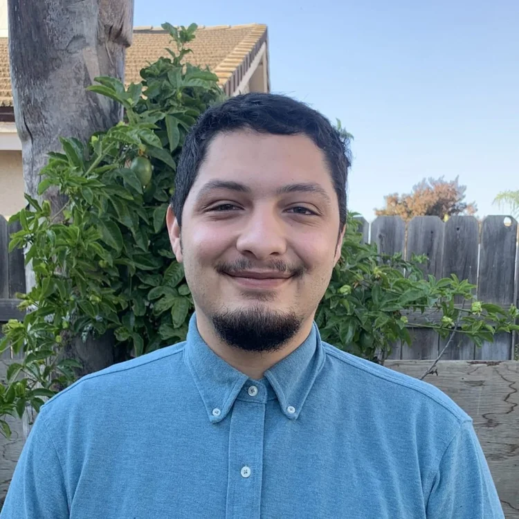 A young man with short dark hair and a goatee is smiling in a backyard with a wooden fence, green foliage, and a house with a sloped roof in the background.