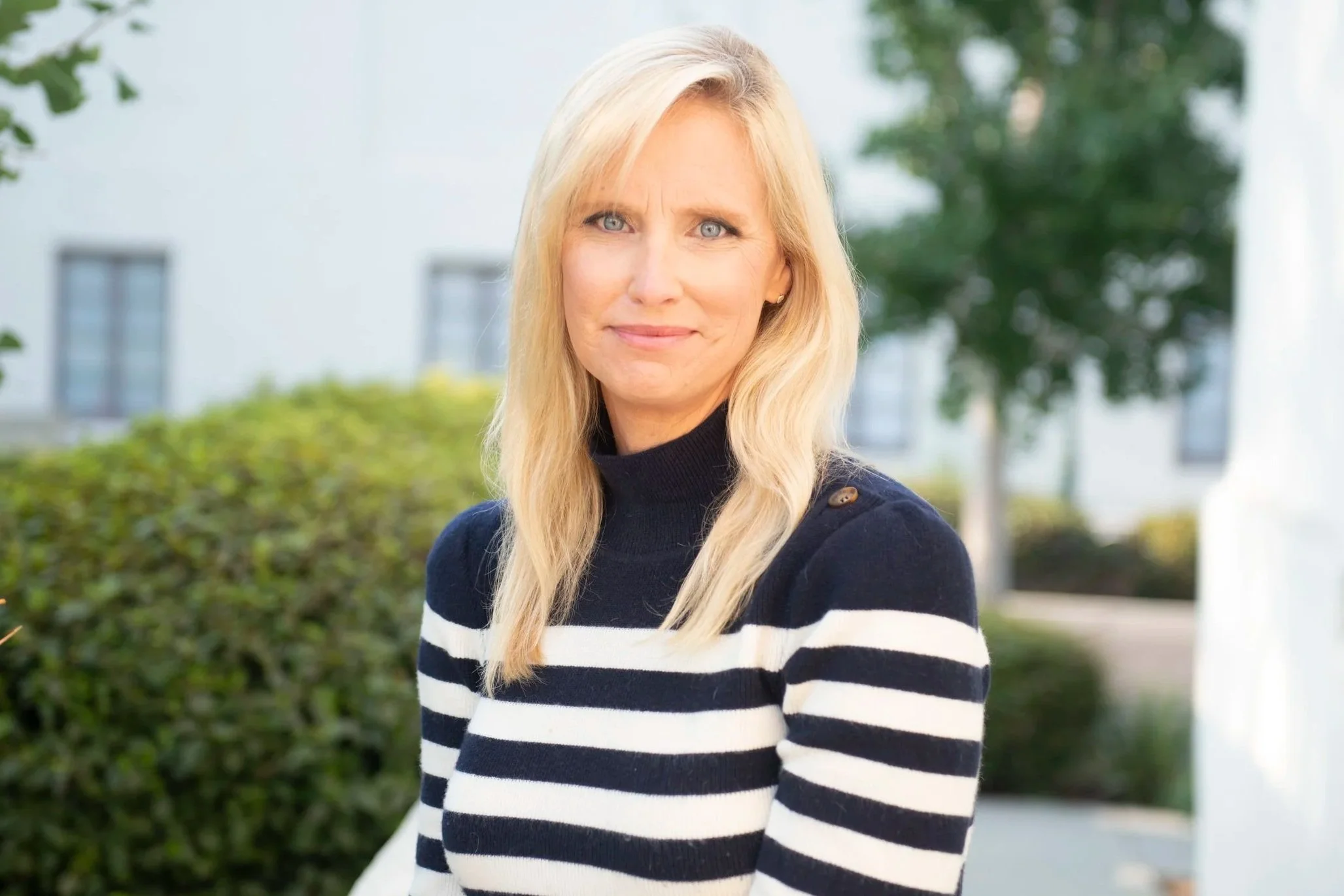 A blonde woman wearing a navy and white striped sweater outdoors, standing in front of bushes and trees with a building in the background.