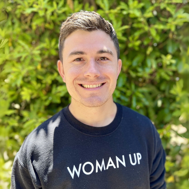 A young man with short brown hair smiling outdoors in front of green foliage, wearing a black sweatshirt that says "WOMAN UP."