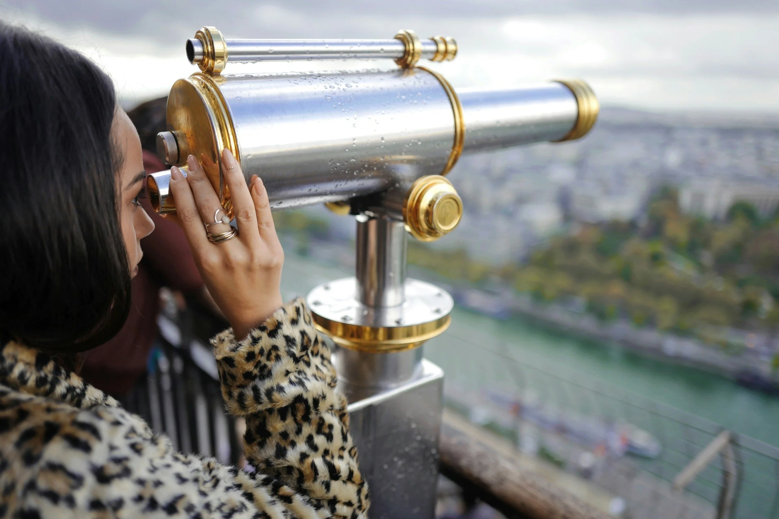 A woman looking through a coin-operated telescope on a city lookout, wearing a leopard print coat.