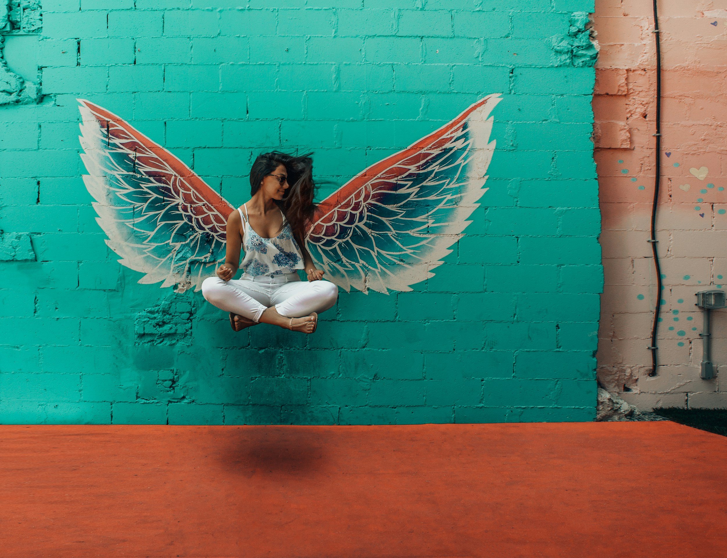 A woman sitting in a cross-legged position with a floating effect, in front of a painted mural of large angel wings on a brick wall.