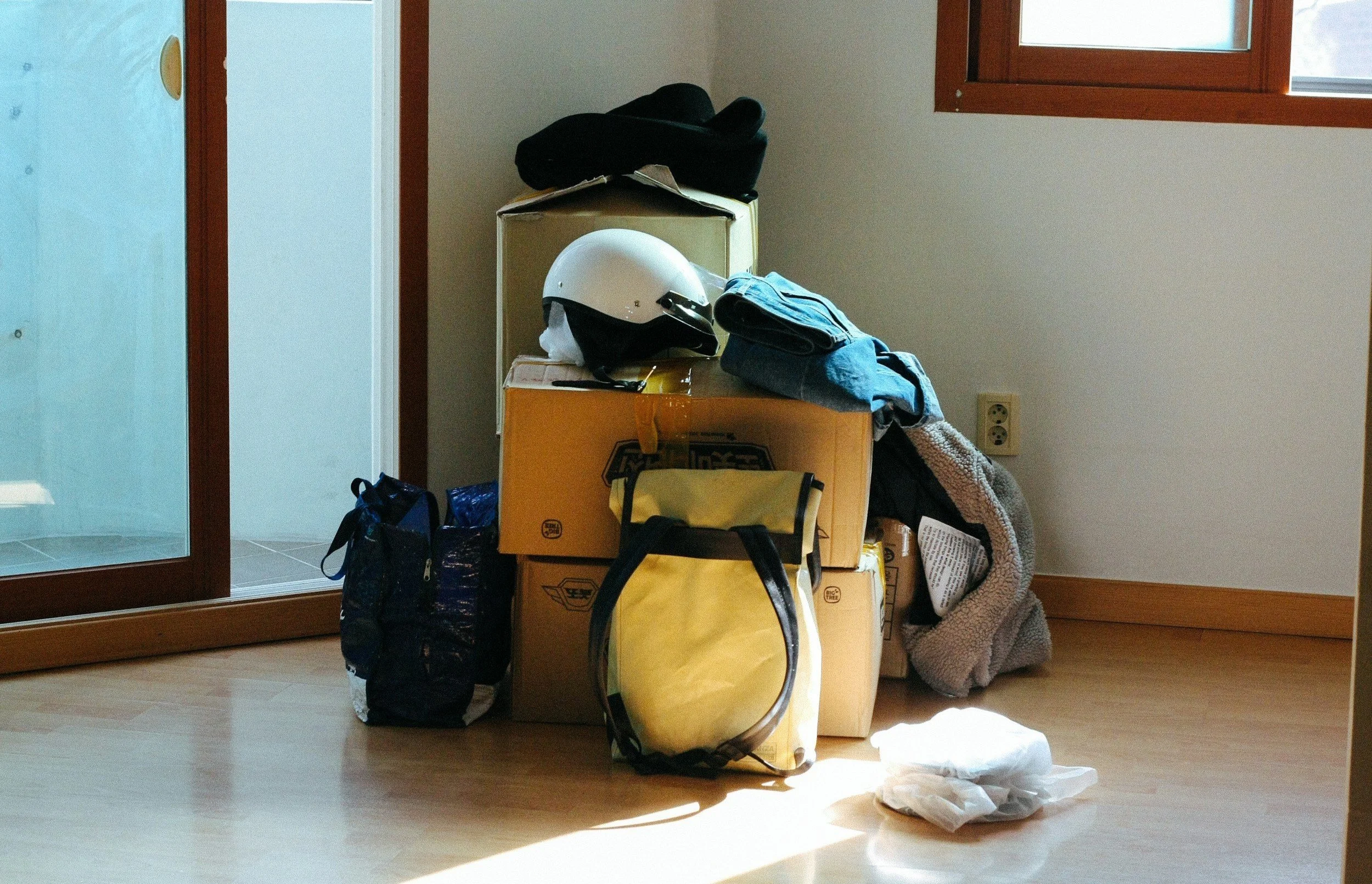 A pile of cardboard boxes with a helmet, clothes, and bags on top, next to a sliding glass door and a window in a room with wooden floors.