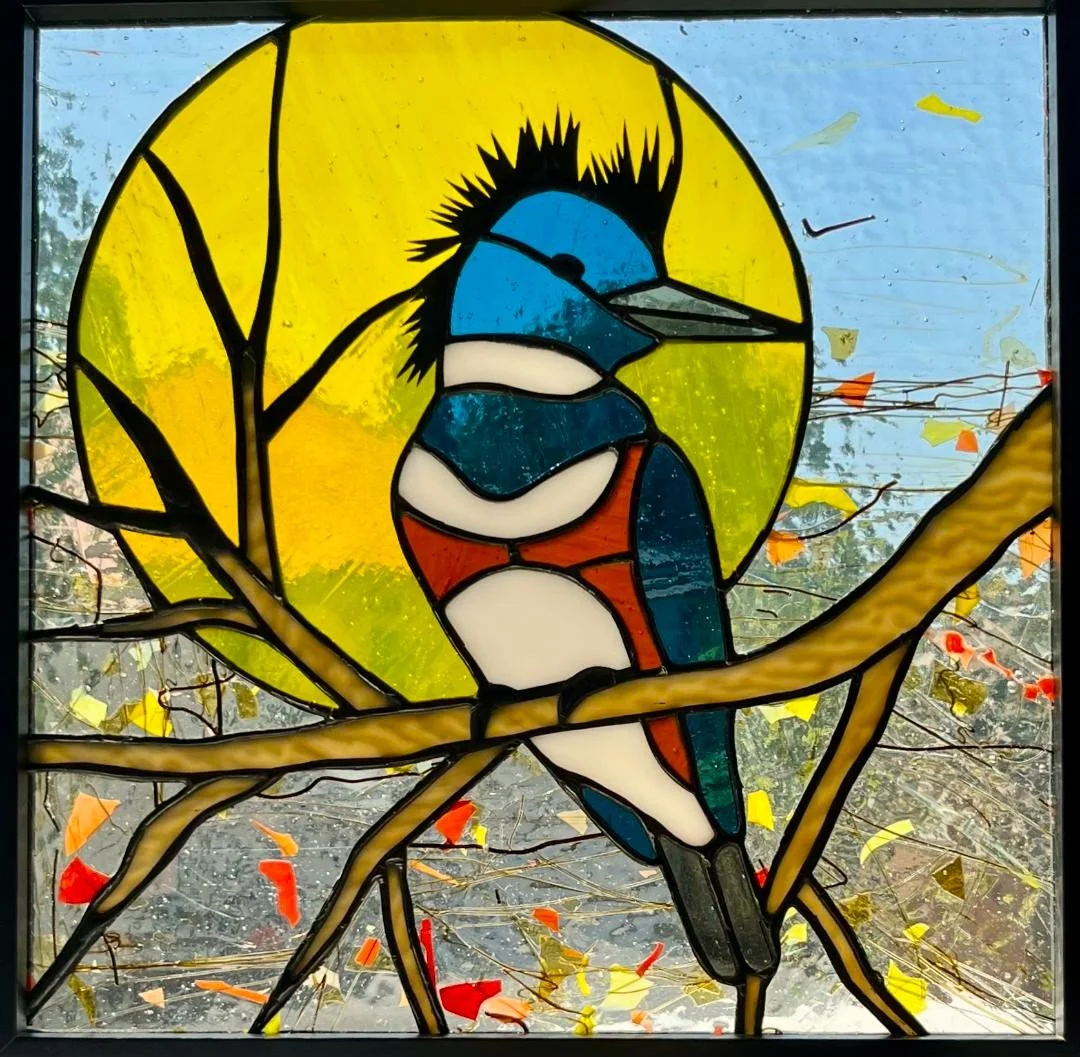 A stained glass window depicting a belted kingfisher bird, perched on a branch with a yellow sun background and orange and red accents.