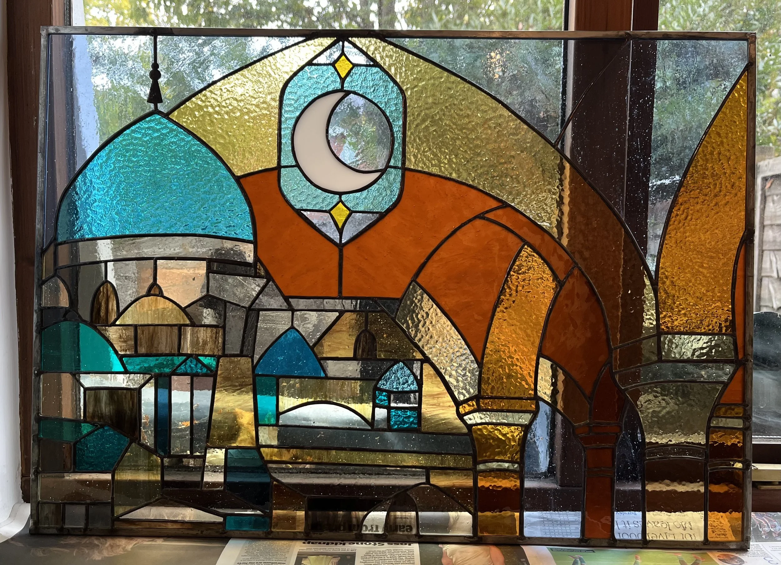 Stained glass window depicting a cityscape with domed buildings, arched doorways, and a crescent moon in a night sky.