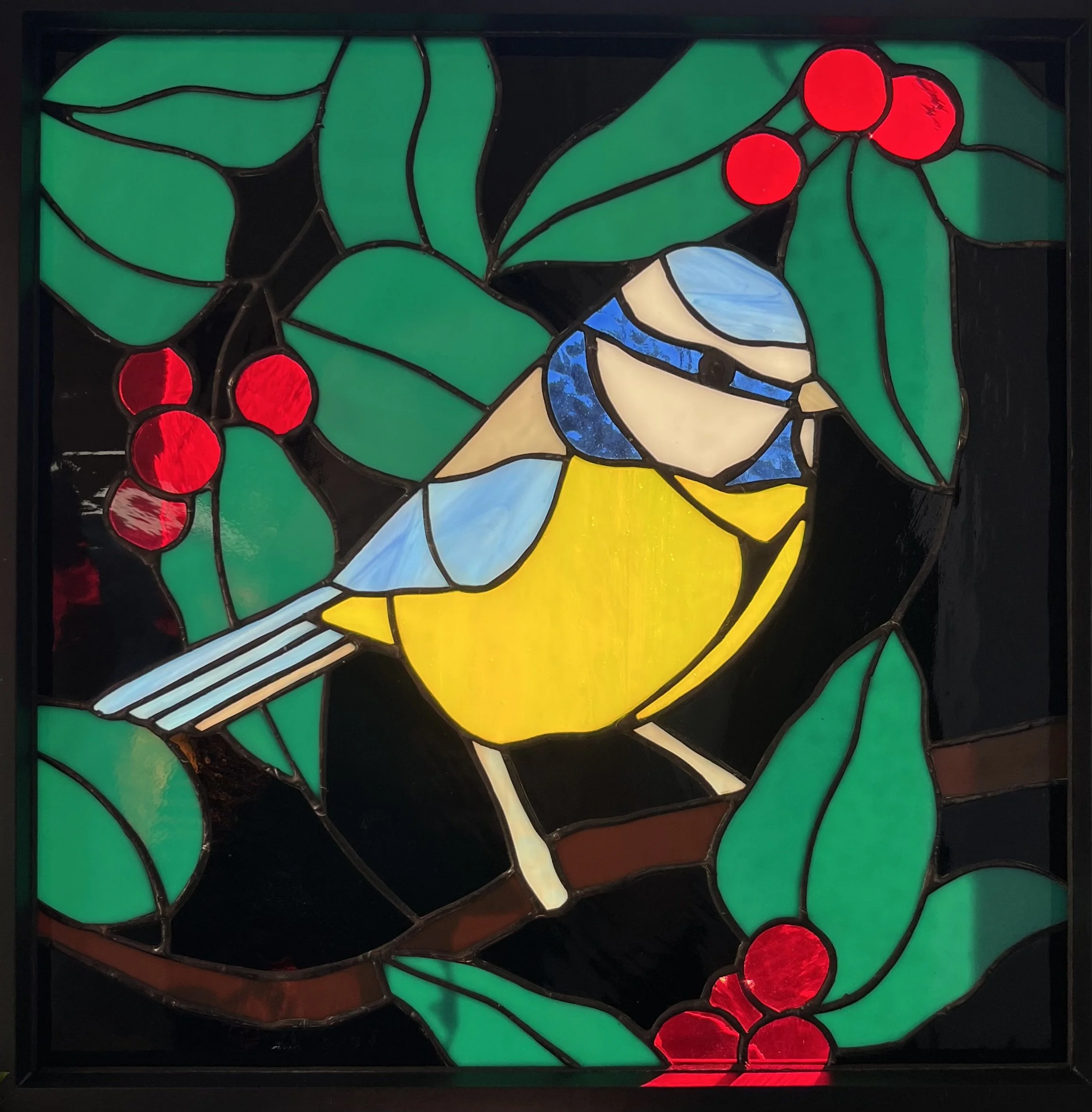 Stained glass window depicting a blue and yellow Blue tit bird perched on a branch among green leaves and red berries.