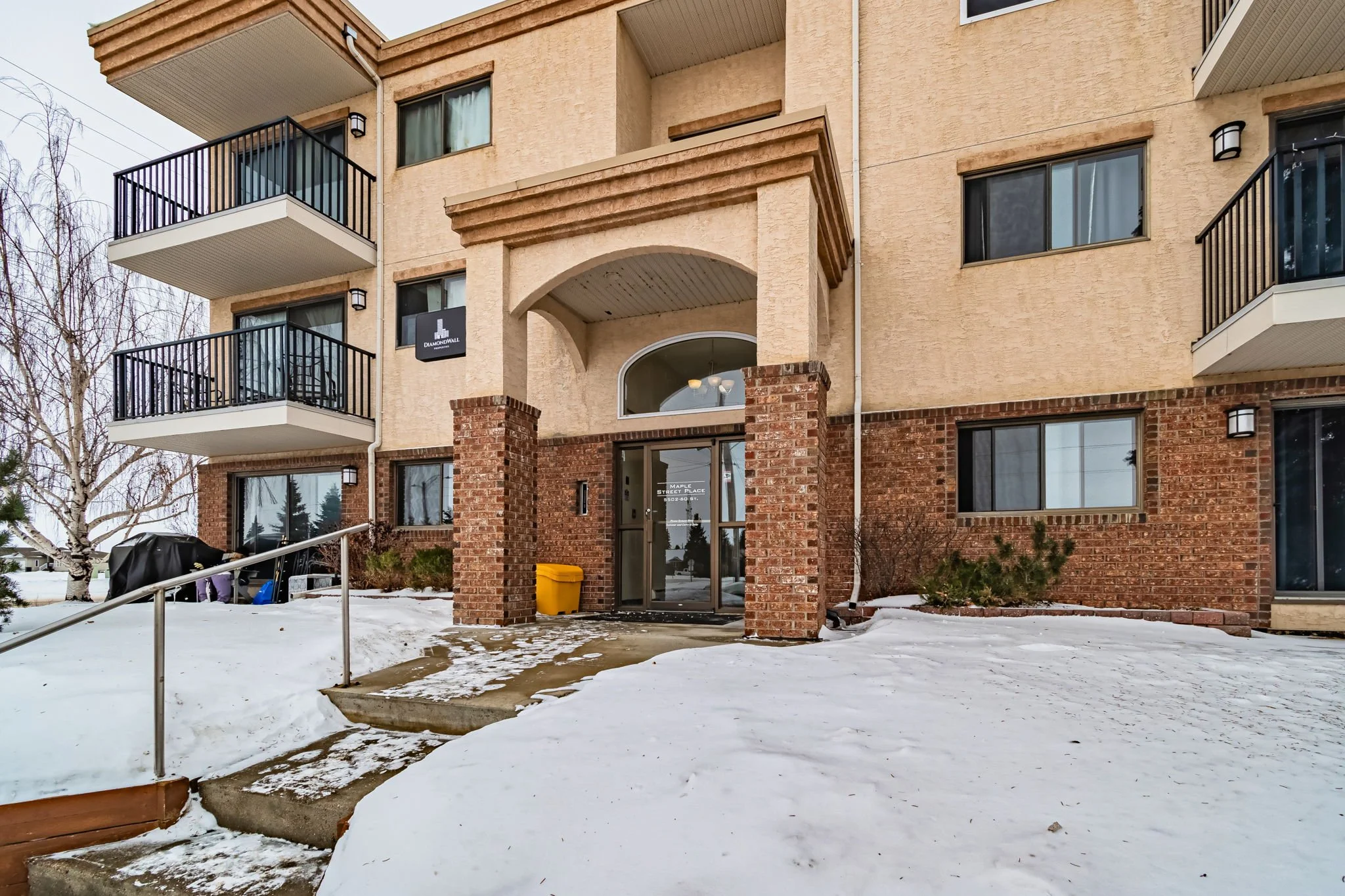 Entrance of a brick apartment building with balconies, surrounded by snow, and a concrete staircase with a metal railing.