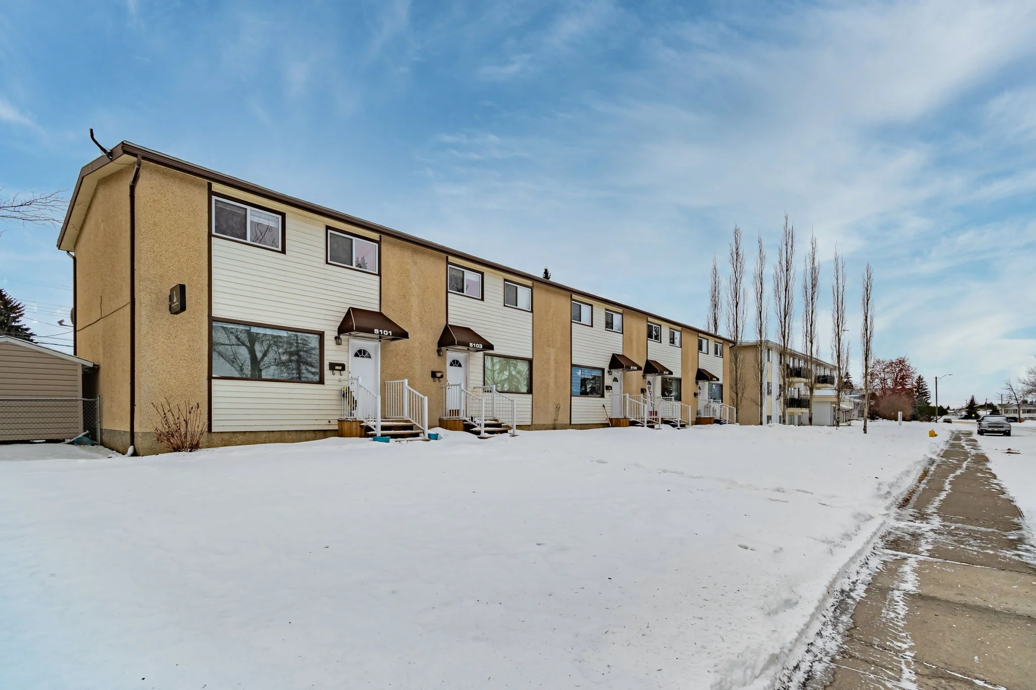 Row of beige townhouses with brown awnings in a snowy landscape, clear sky above.