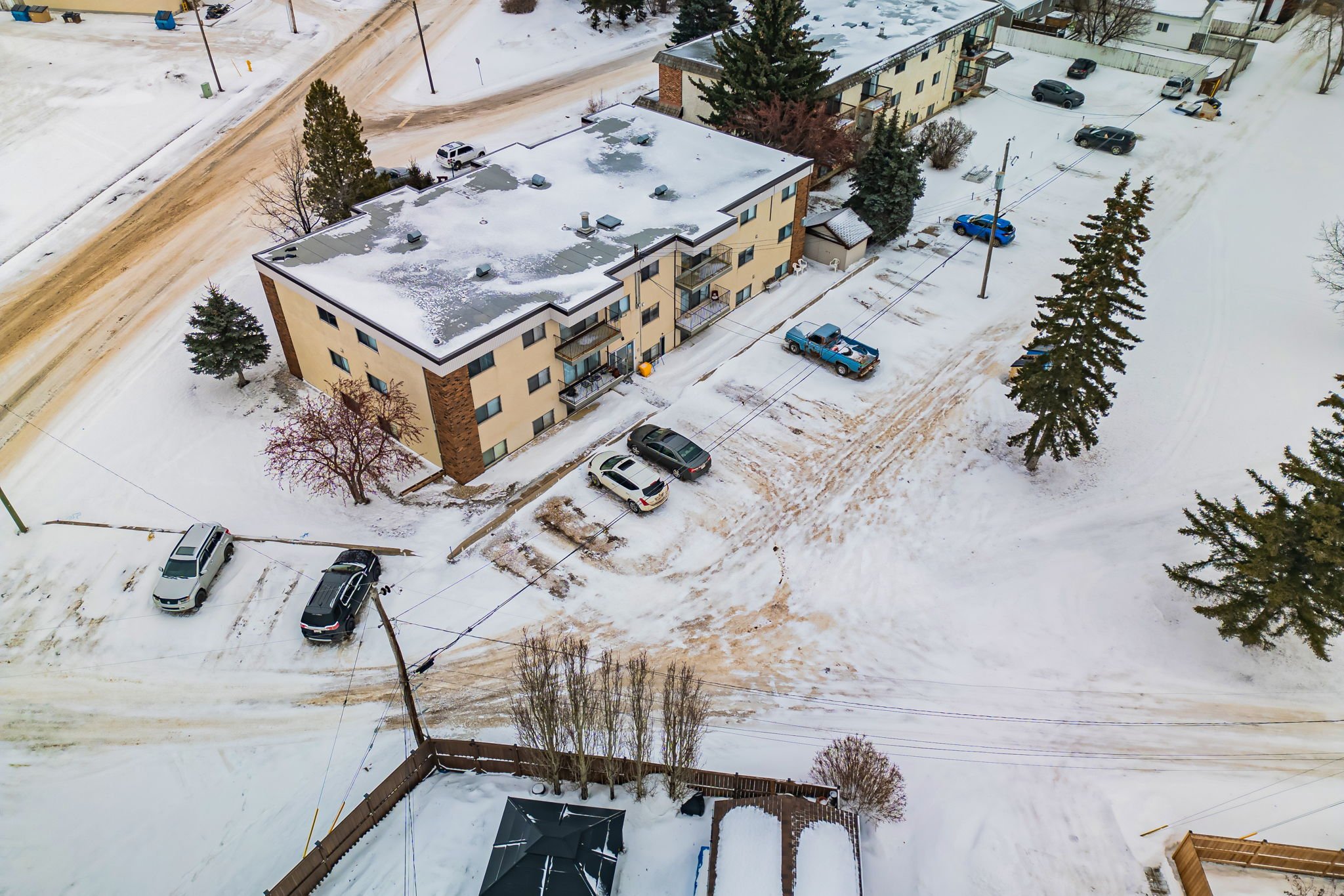 Aerial view of snow-covered street and apartment buildings with parked cars and trees.