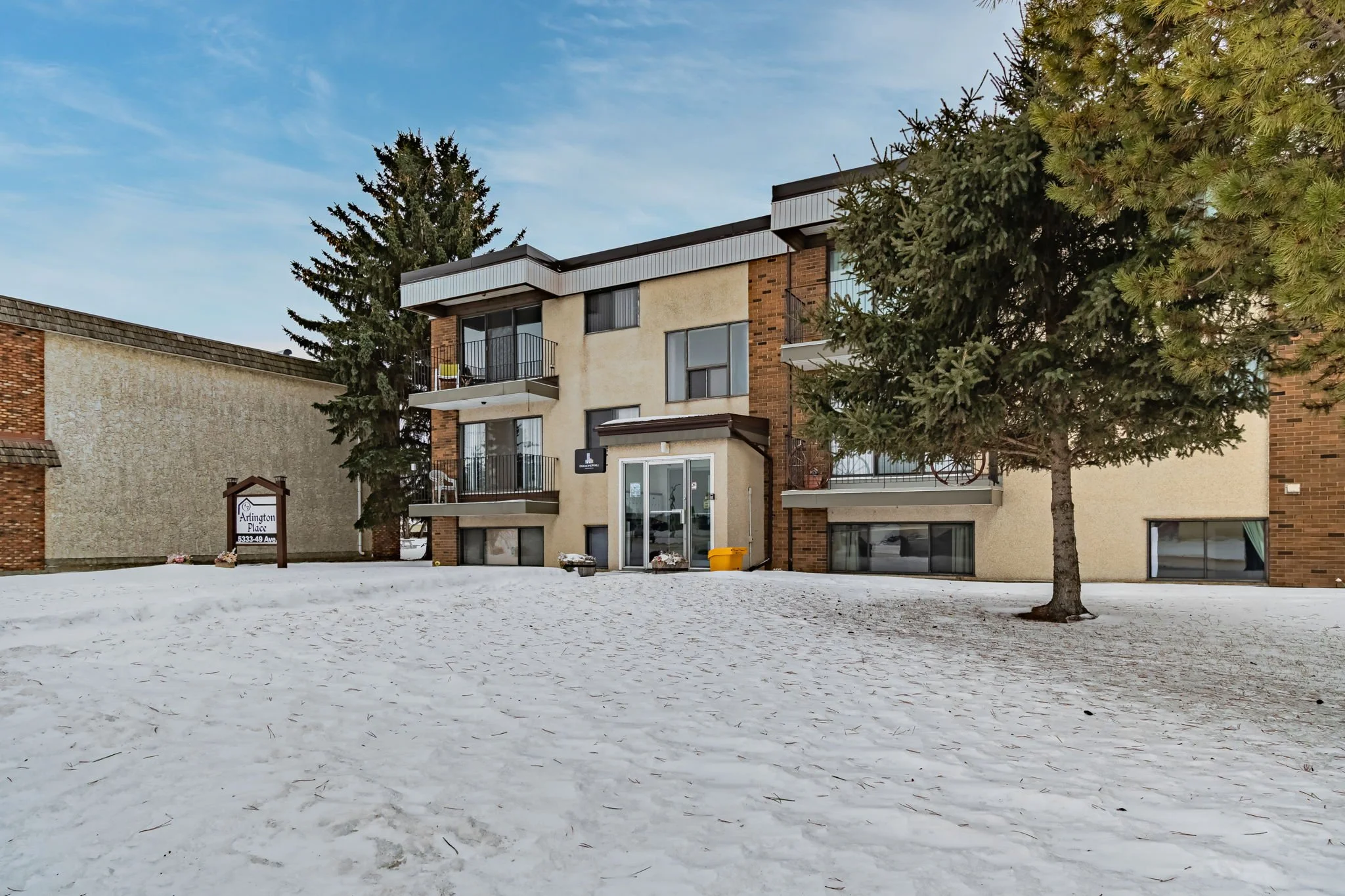 Three-story residential apartment building with balconies in a snowy landscape, surrounded by trees, labeled "Arlington Place."