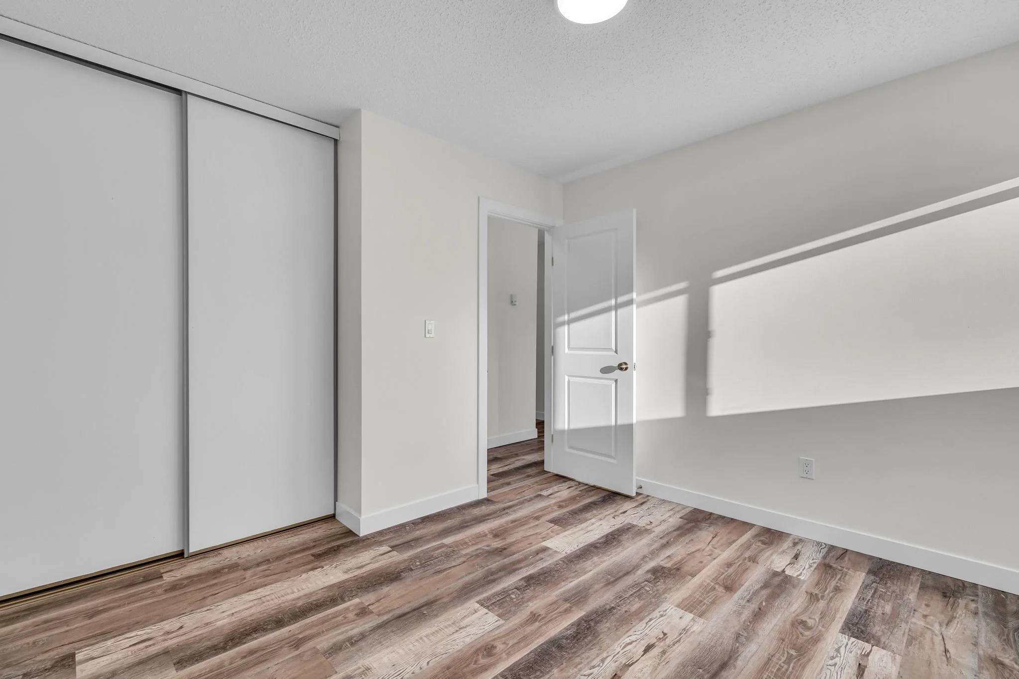 Empty room with wood laminate flooring, light-colored walls, a white door partially open, and sliding closet doors.