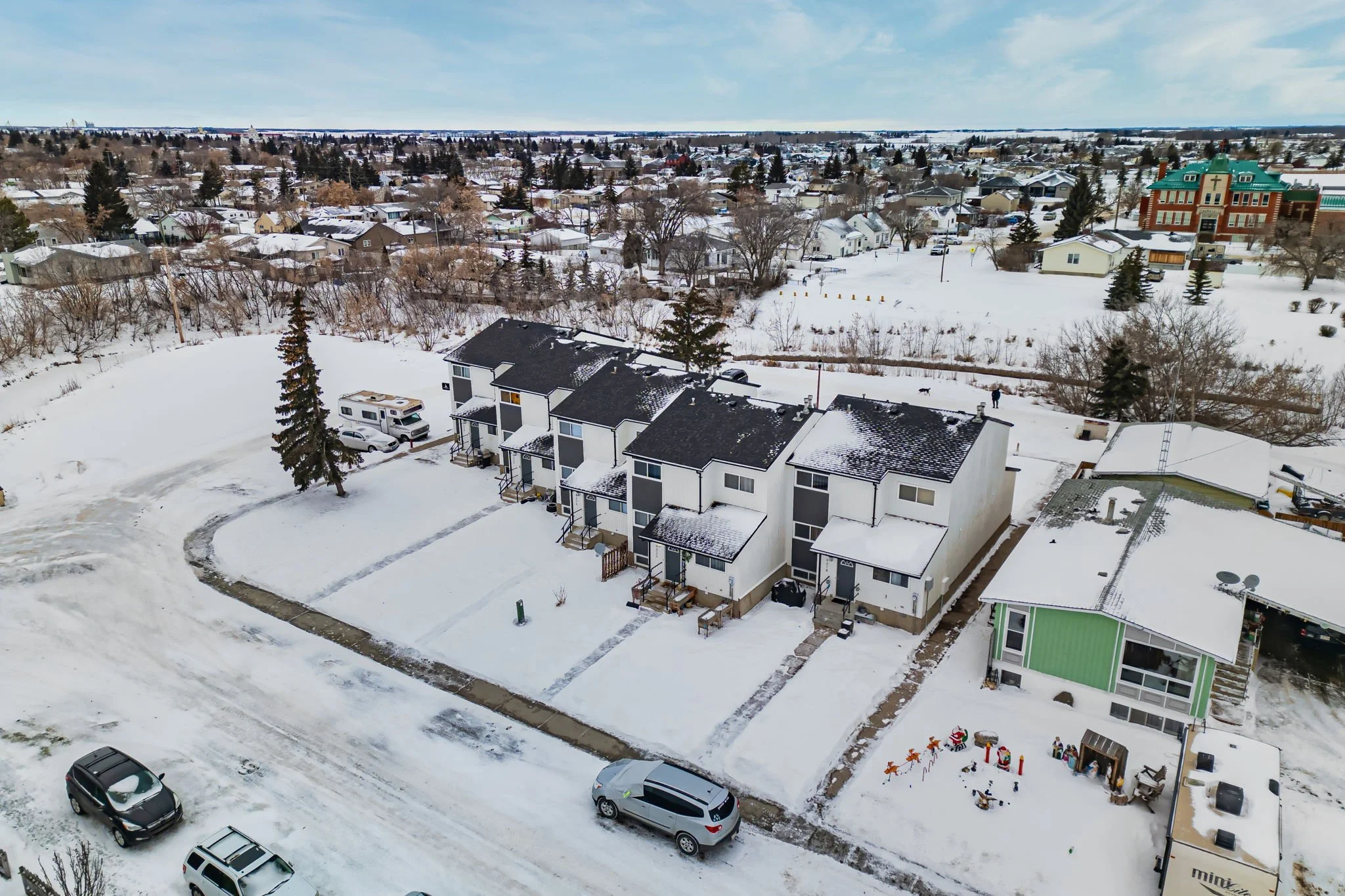 Aerial view of snow-covered residential area with a row of contemporary townhouses surrounded by roads and parked cars, adjacent to a single-story building and scattered trees.