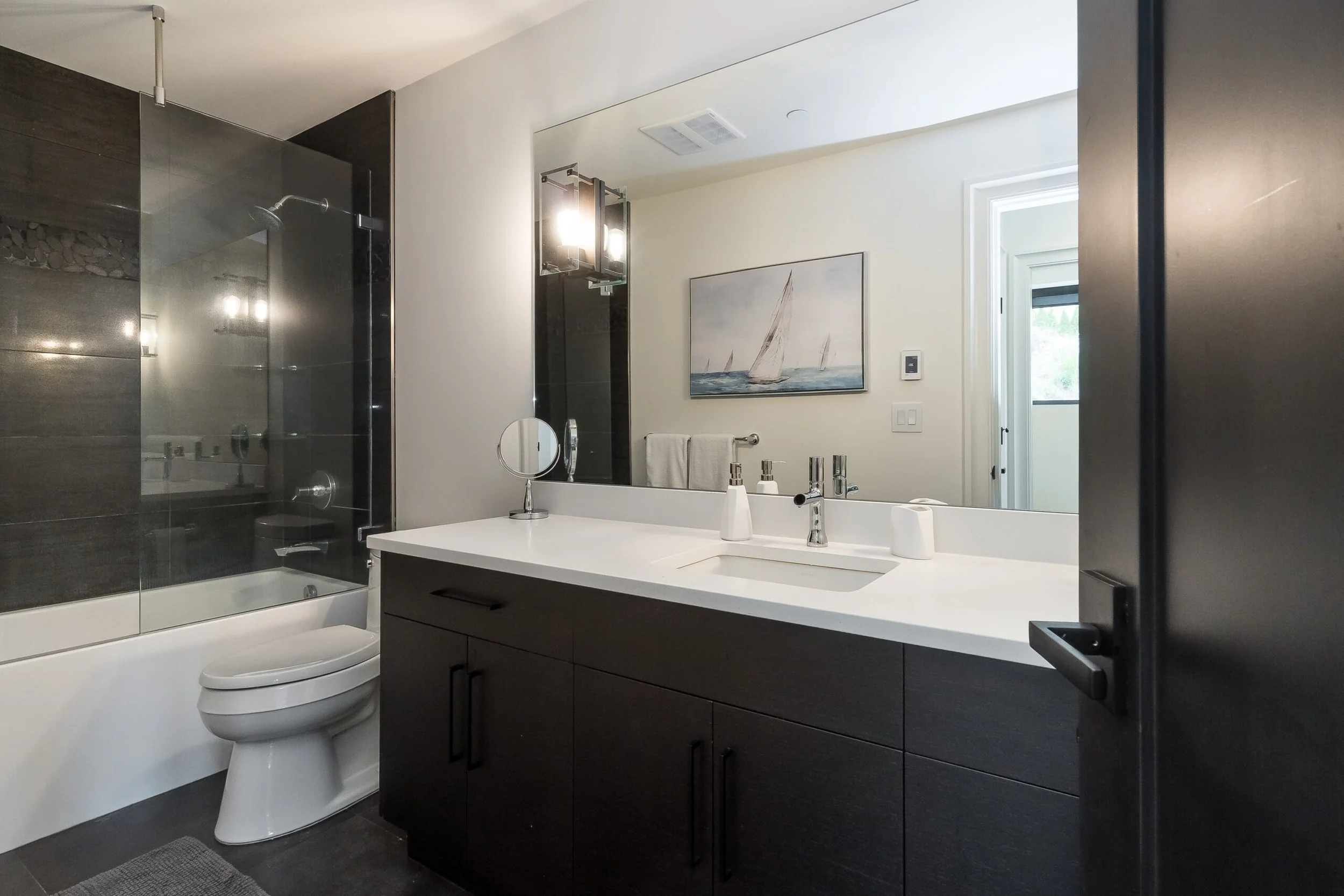 Modern bathroom with a vanity, sink, large mirror, toilet, bathtub, and glass shower enclosure, featuring dark cabinetry and a sailboat painting on the wall.