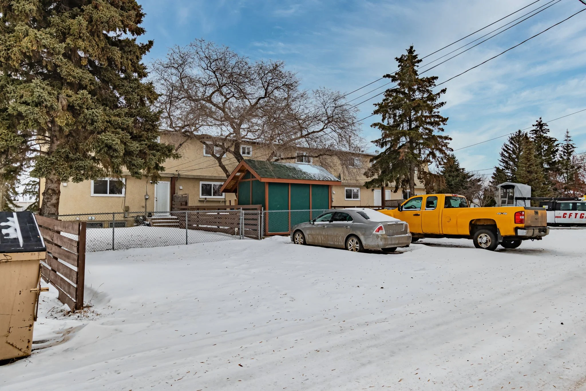 Snowy residential area with a beige house, two parked vehicles (a silver car and a yellow truck), large trees, and a wooden fence.