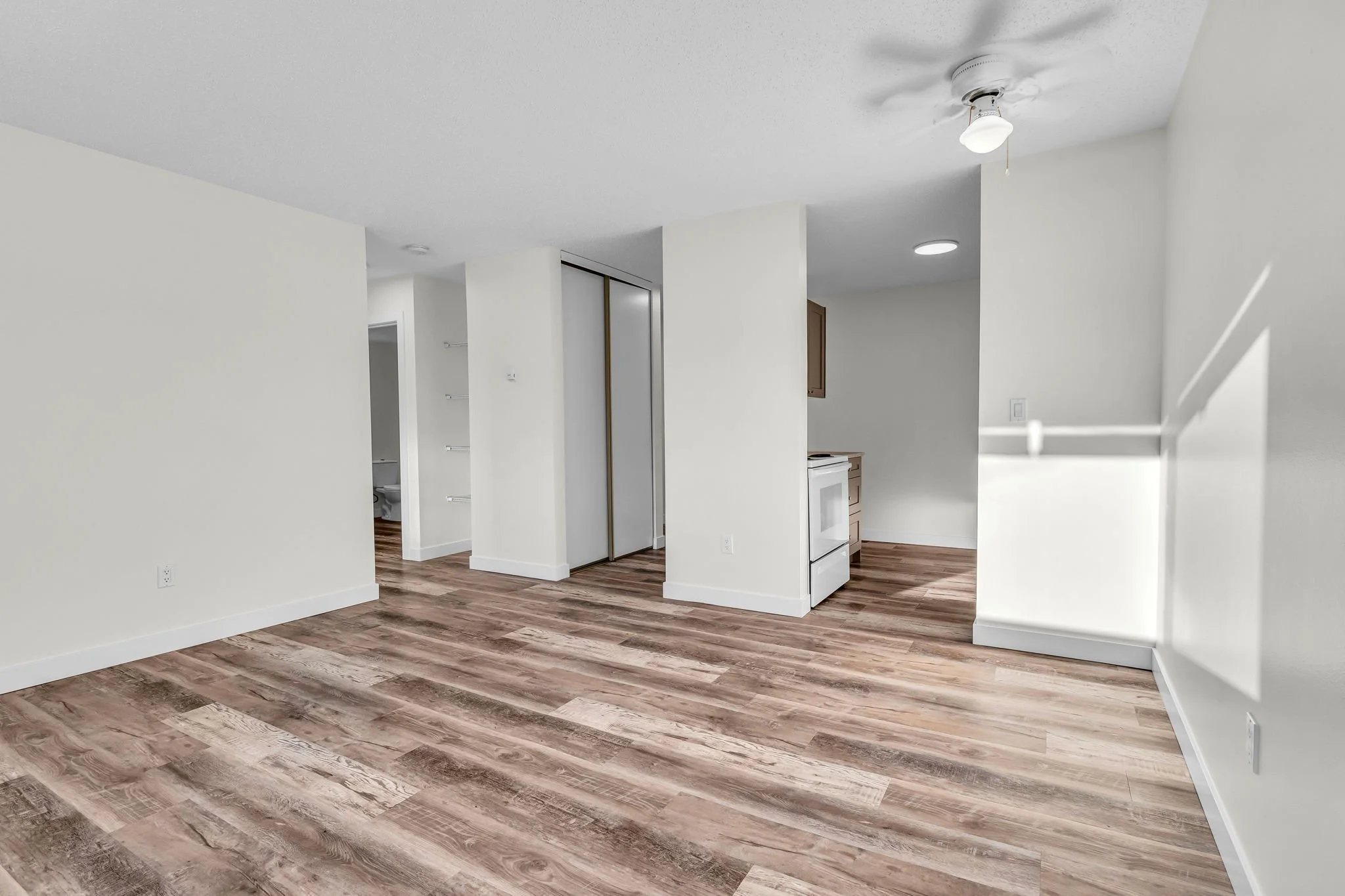 Empty room with wood flooring, light-colored walls, a ceiling fan, and an open doorway leading to a kitchen with a white stove.