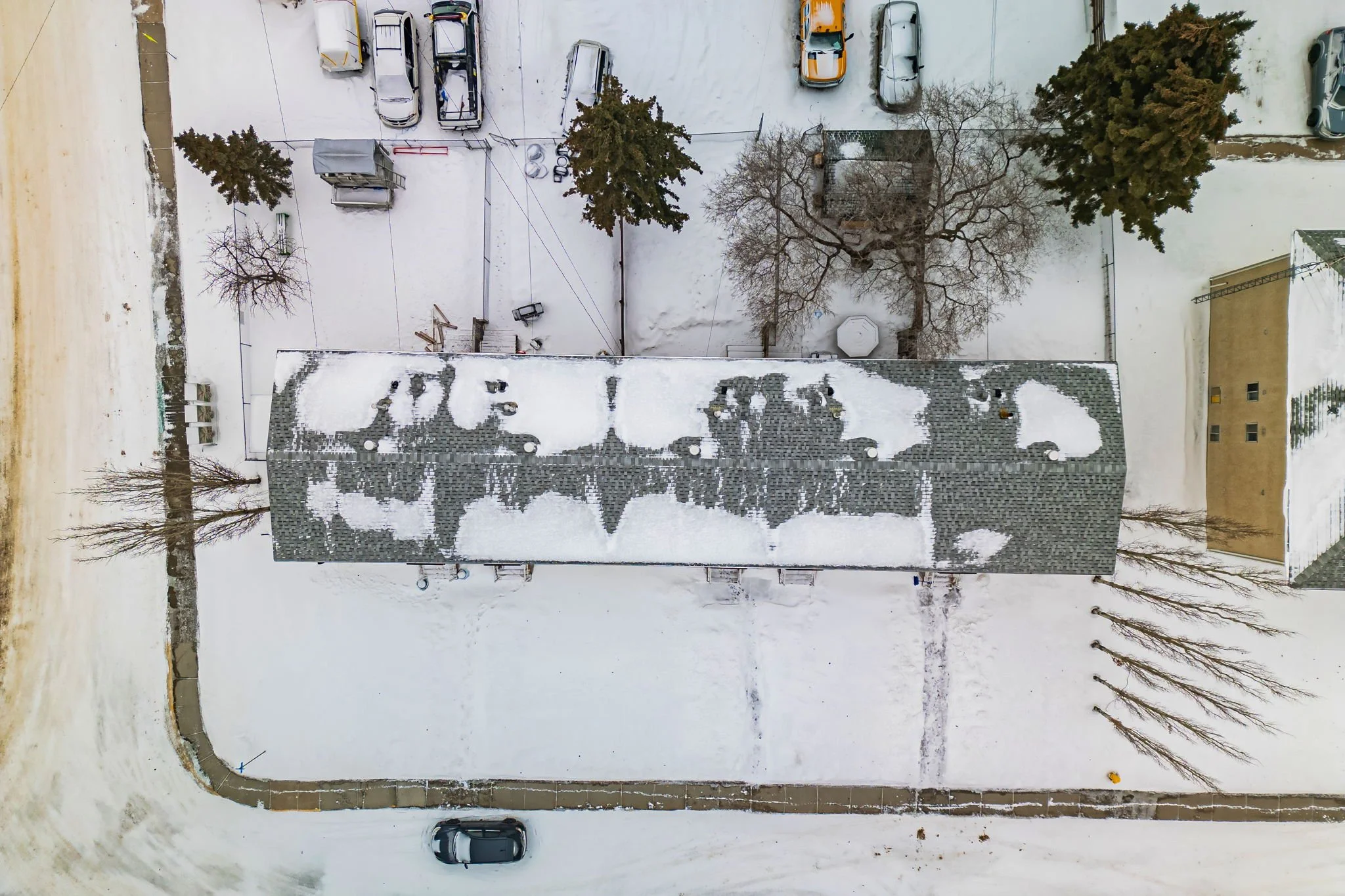 Aerial view of a snow-covered building, parking lot with cars, and surrounding trees in a residential area.