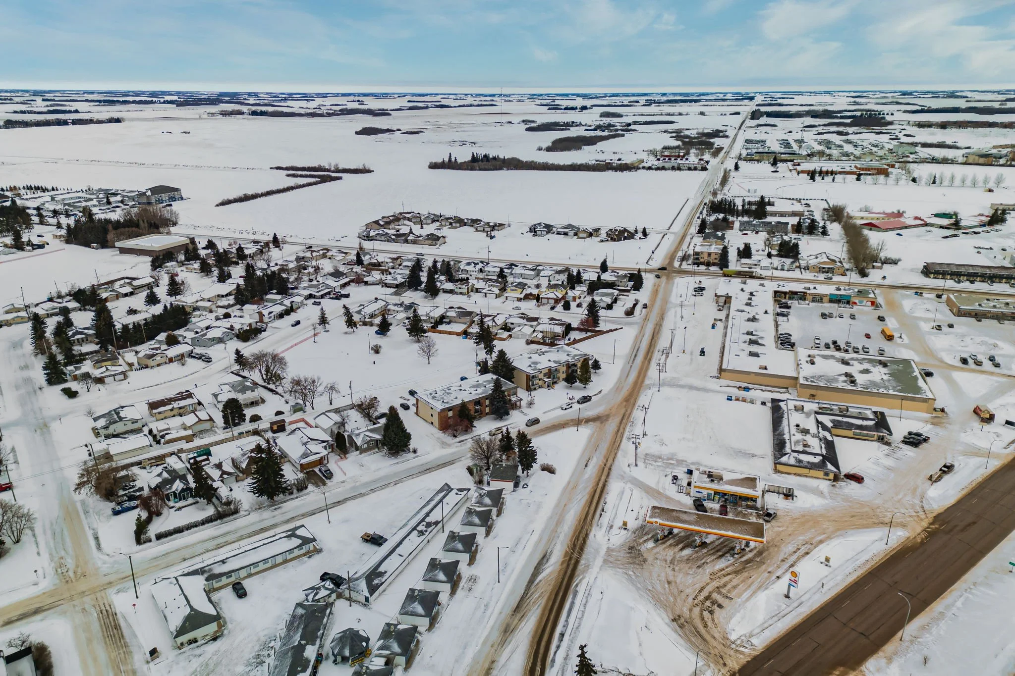 Aerial view of a snowy rural town with roads, houses, a gas station, and fields in the background.