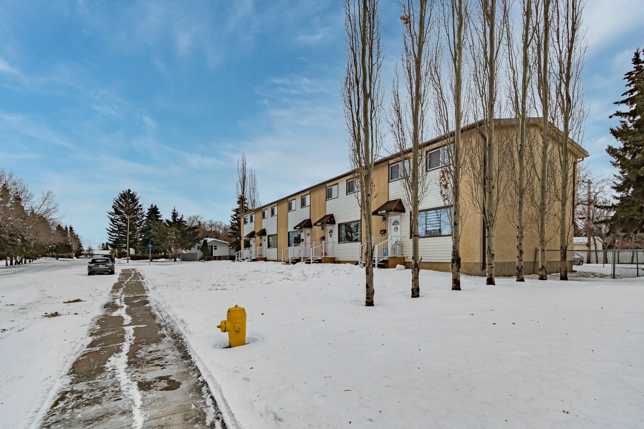Snow-covered suburban street with townhouses, trees, a yellow fire hydrant, and a parked car.