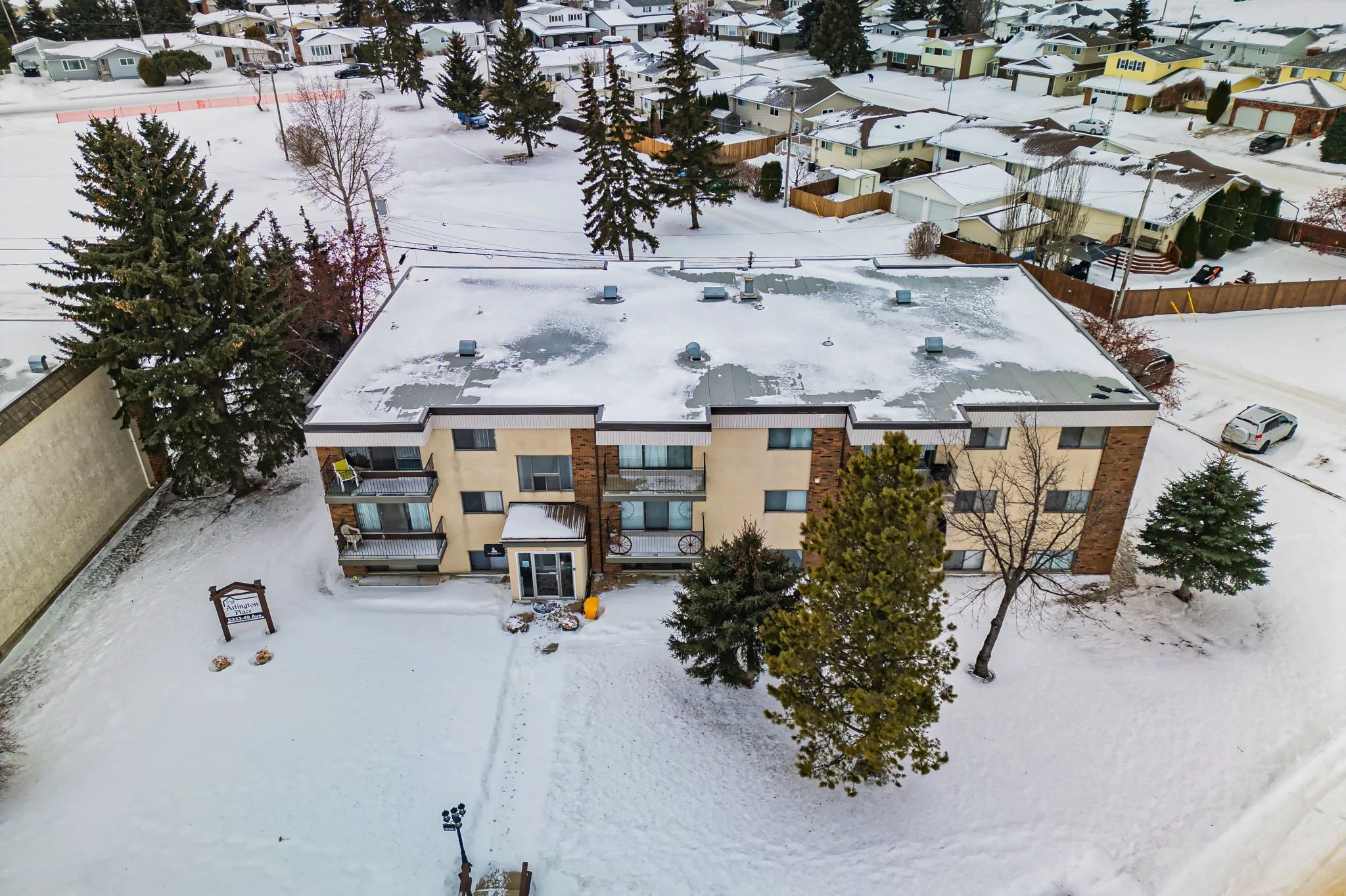 Aerial view of a snow-covered apartment building with surrounding trees and a parking lot, during winter in a residential neighborhood.