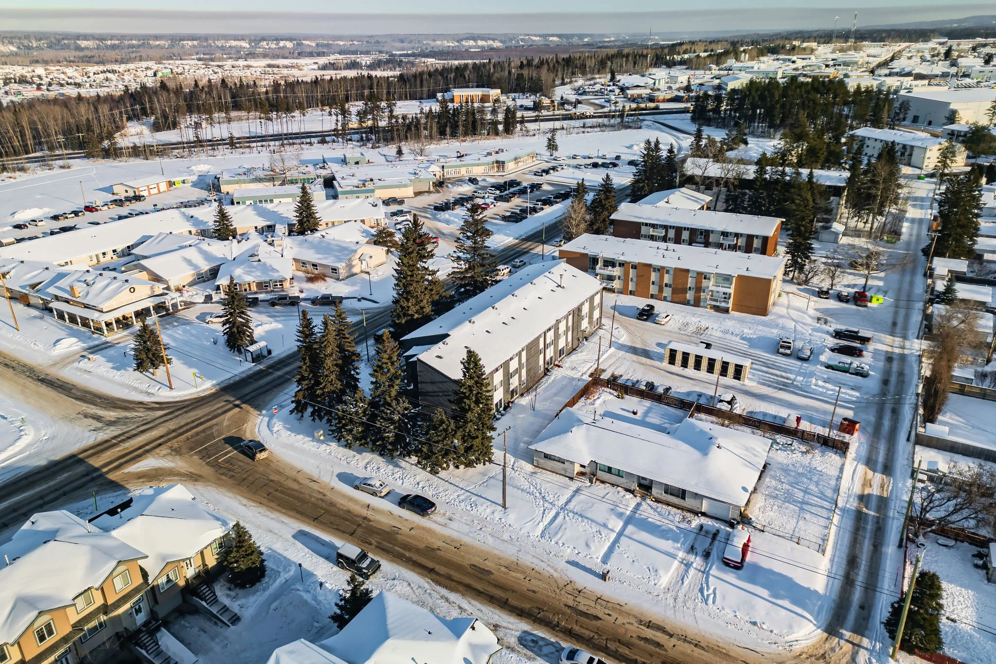Aerial view of a snowy suburban area with residential buildings, roads, and trees. The landscape is covered in snow and includes a mix of houses and apartment buildings. Cars are parked along the streets, and evergreen trees are visible throughout the scene.