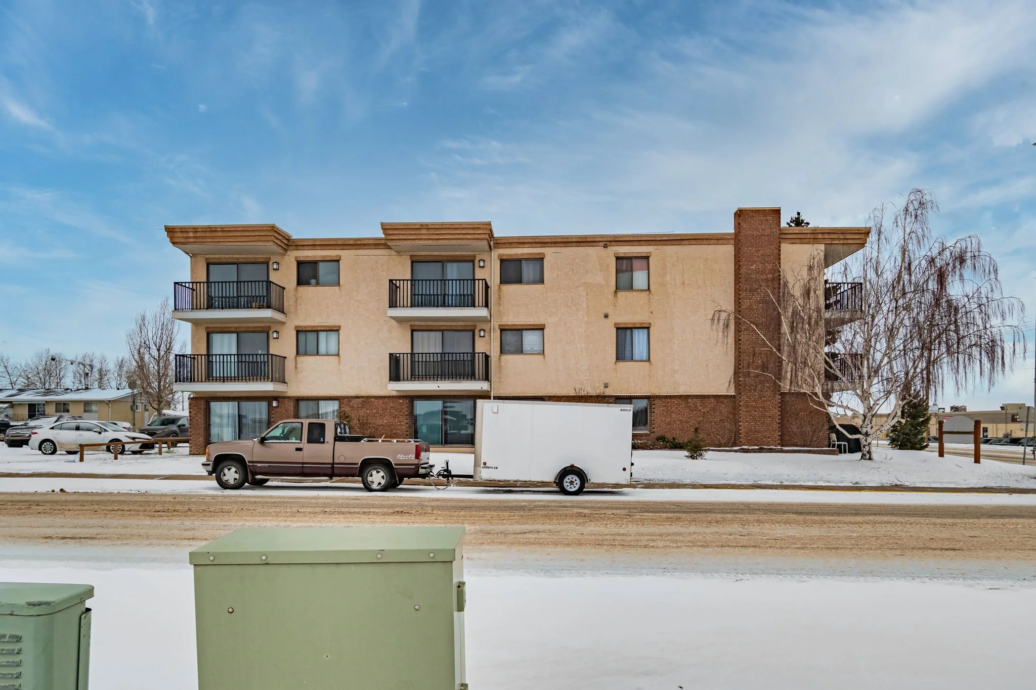Three-story apartment building with balconies, parked truck and trailer, winter snow on the ground, overcast sky.