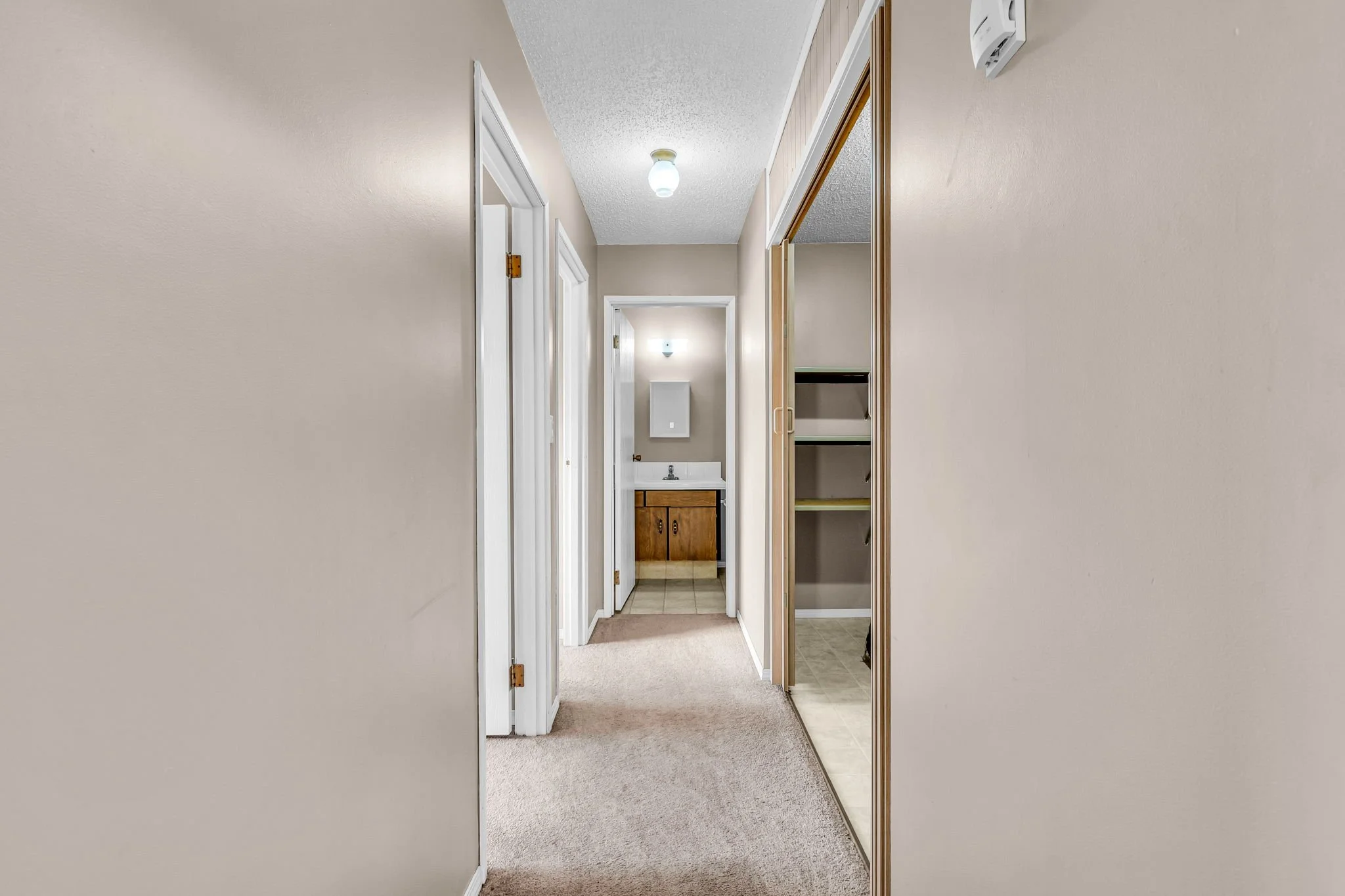 Interior hallway with beige walls, carpeted floor, closet with sliding doors, and view into a bathroom.