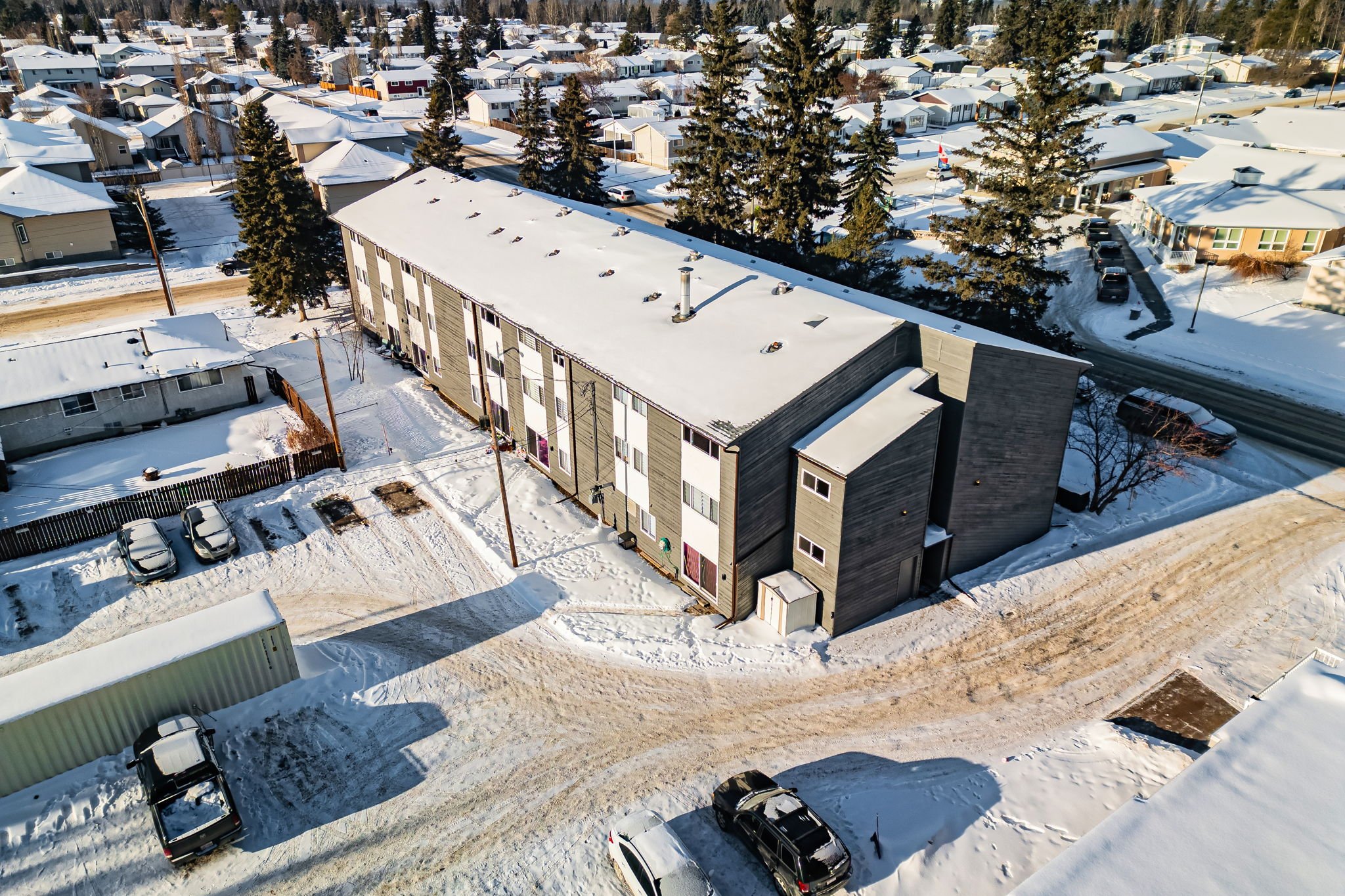 Aerial view of an apartment building and surrounding residential area covered in snow, with parked cars and trees.