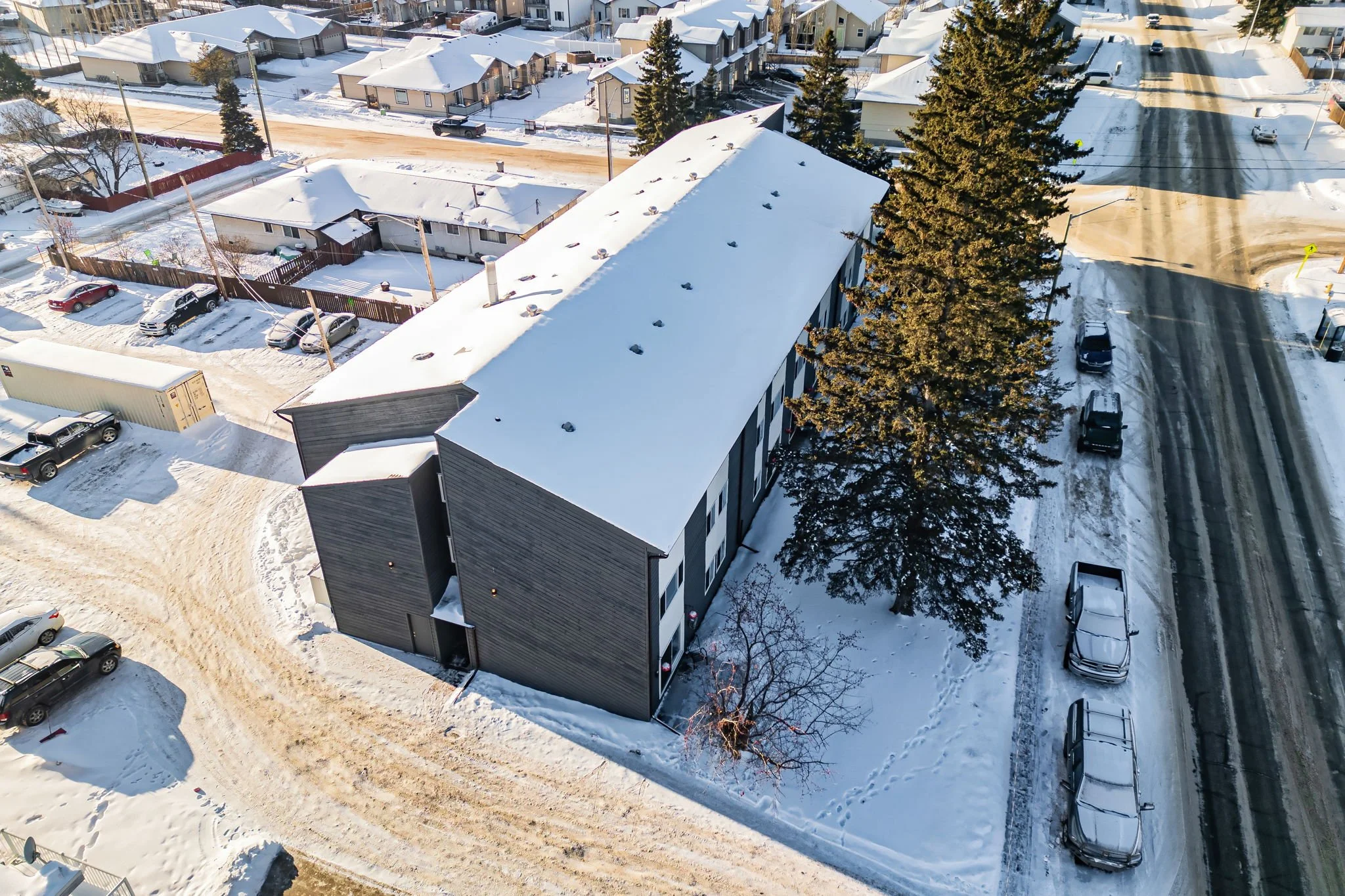 Aerial view of a snowy residential neighborhood with parked cars and a long building, surrounded by trees and roads.
