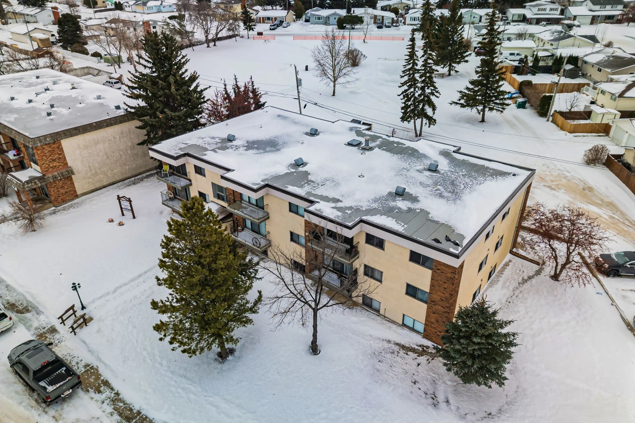 Aerial view of a snow-covered residential building with surrounding trees, parked vehicles, and adjacent neighborhood houses.