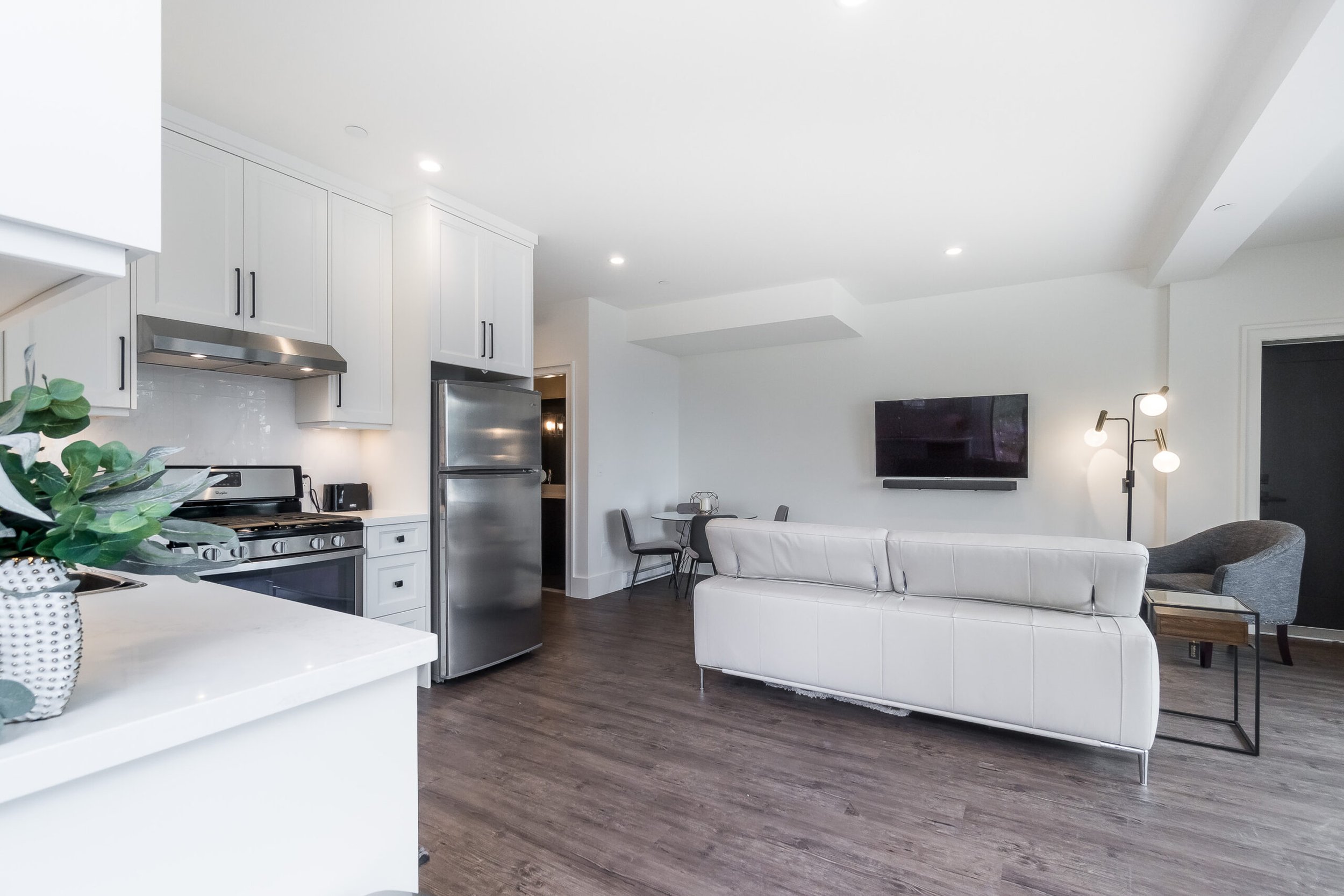 Modern open-concept kitchen and living room with white cabinetry, stainless steel appliances, white sofa, wall-mounted TV, and wooden flooring.