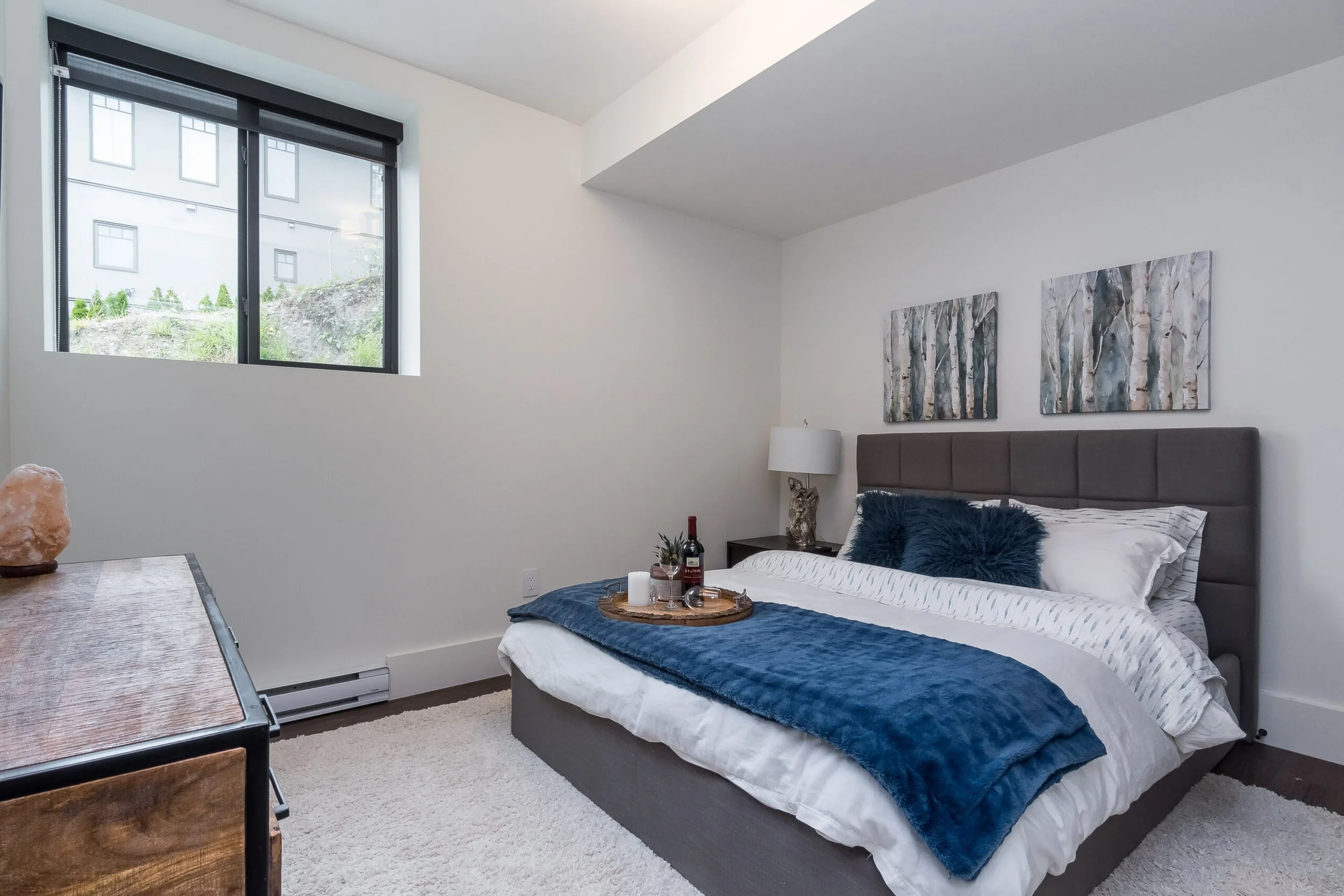 Modern bedroom with a large bed, blue blanket, and decorative pillows, two abstract paintings above the headboard, side table with a lamp, and a window with a view outside.