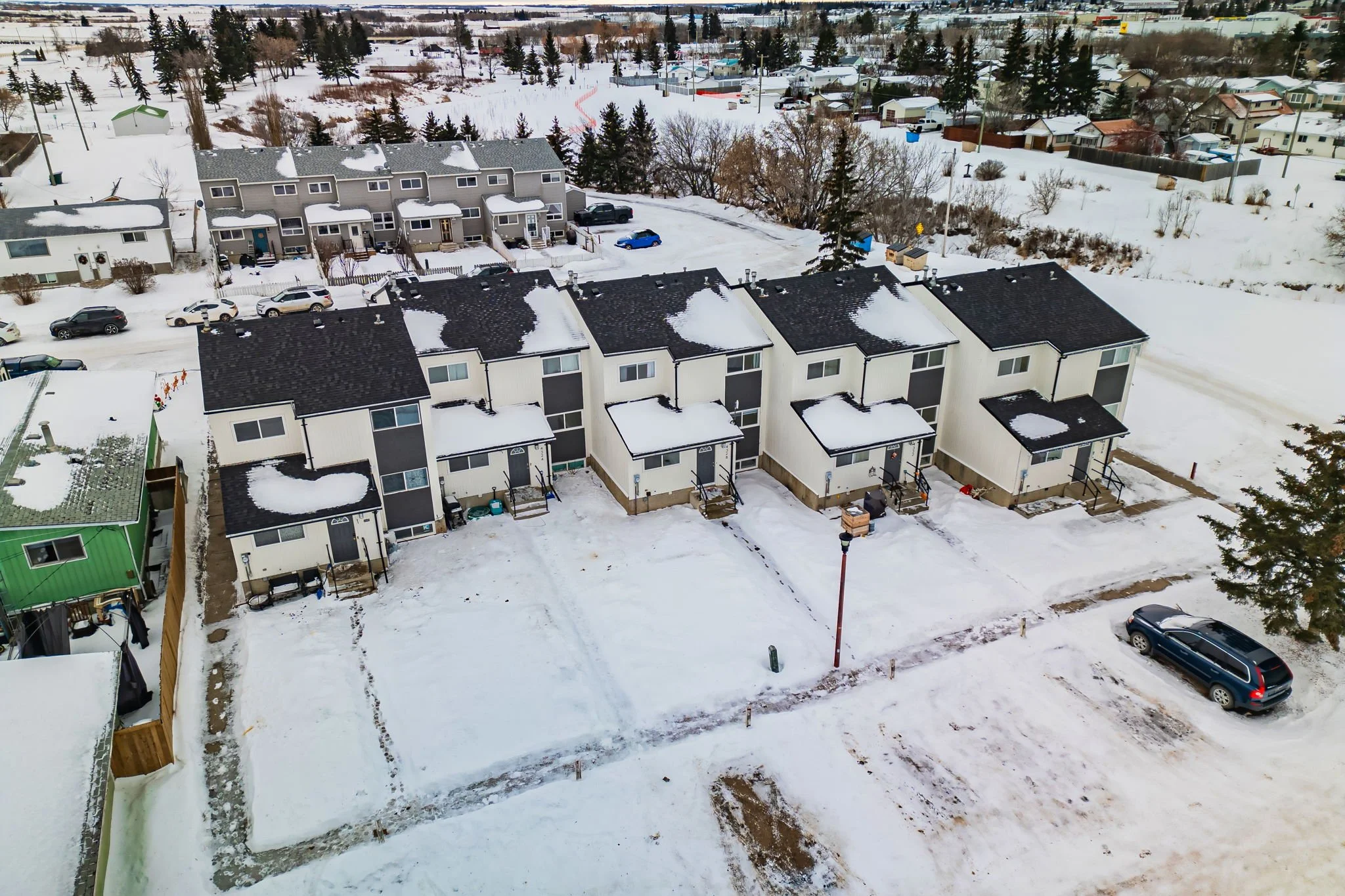 Aerial view of a snowy residential neighborhood featuring modern townhouses with dark roofs, surrounded by cars and snow-covered roads.