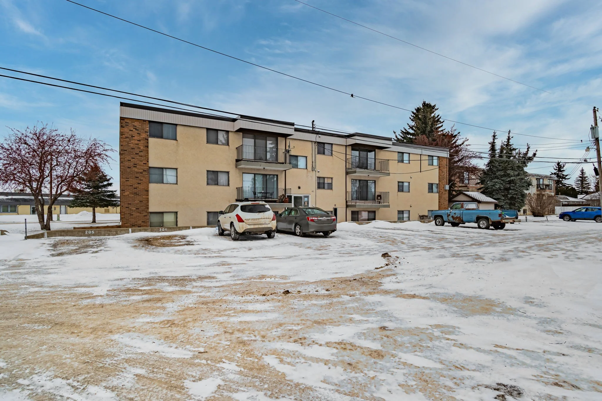 Apartment building with snowy parking lot and cars, overcast sky.